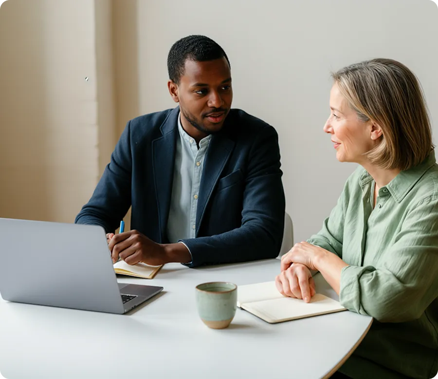 Man and woman having a discussion across a table with a laptop, notebooks, and a mug.