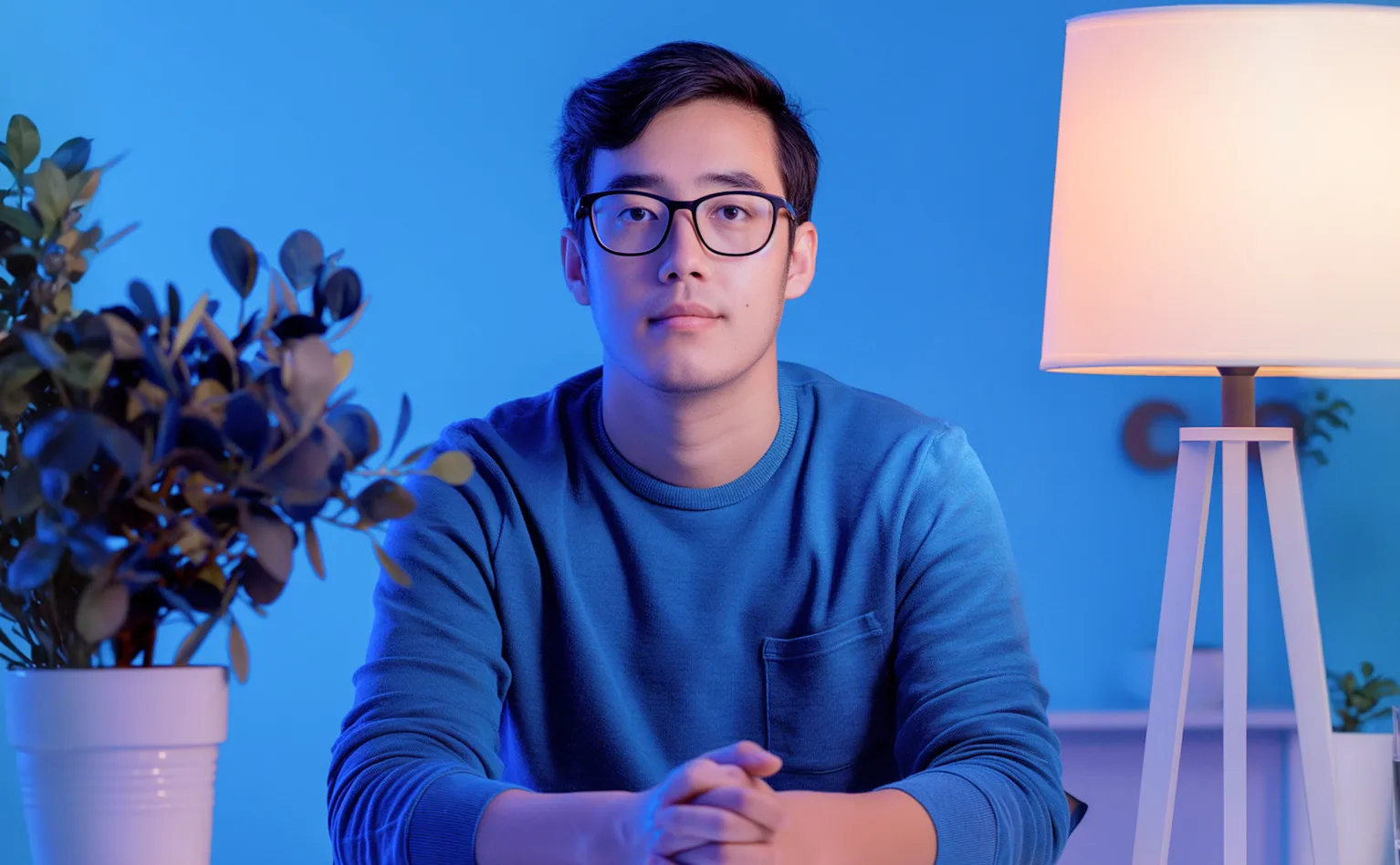 Young man wearing glasses and a blue sweater sitting with hands clasped at a table, with a potted plant and a lit lamp in a blue-lit room.