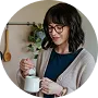 Smiling woman wearing glasses pouring coffee from a French press into a white mug in a kitchen.