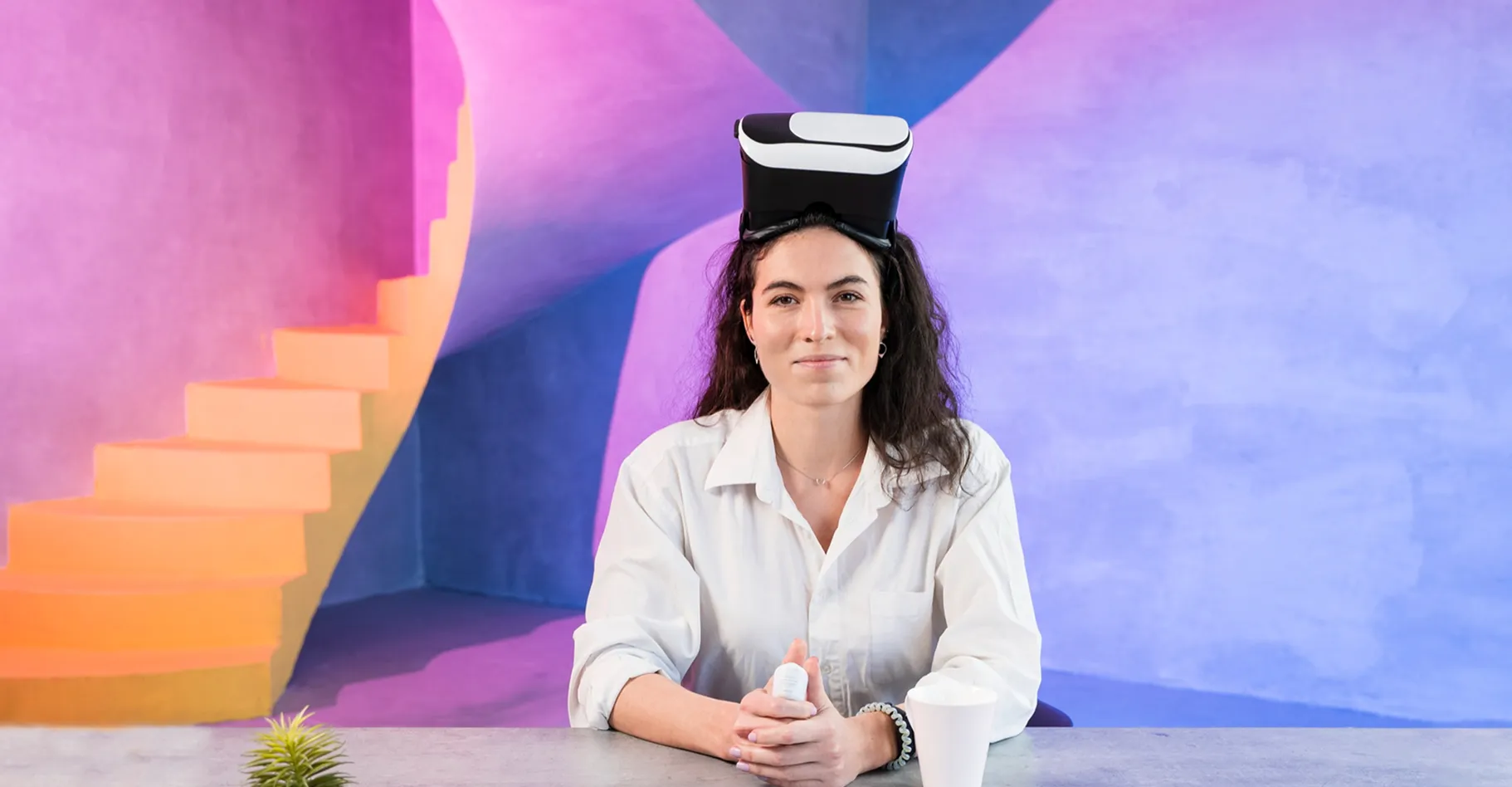 Woman in a white shirt sitting at a table with a virtual reality headset on her head, colorful modern stairs in the background.