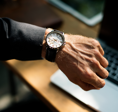 Close-up of a man's wrist wearing a classic analog wristwatch with a leather strap over a desk with a laptop.