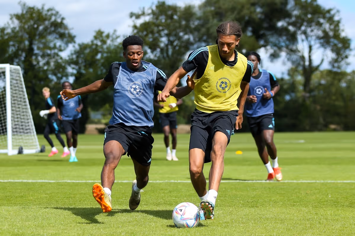 Two professional football  players in training vests competing for the ball on a grassy field with teammates and goalpost in the background.
