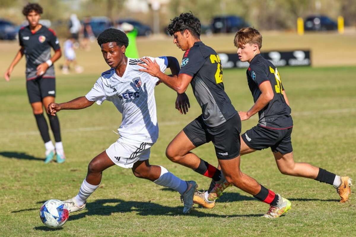 Three young soccer players in action on a grass field, with one player in white controlling the ball while two players in black attempt to defend.