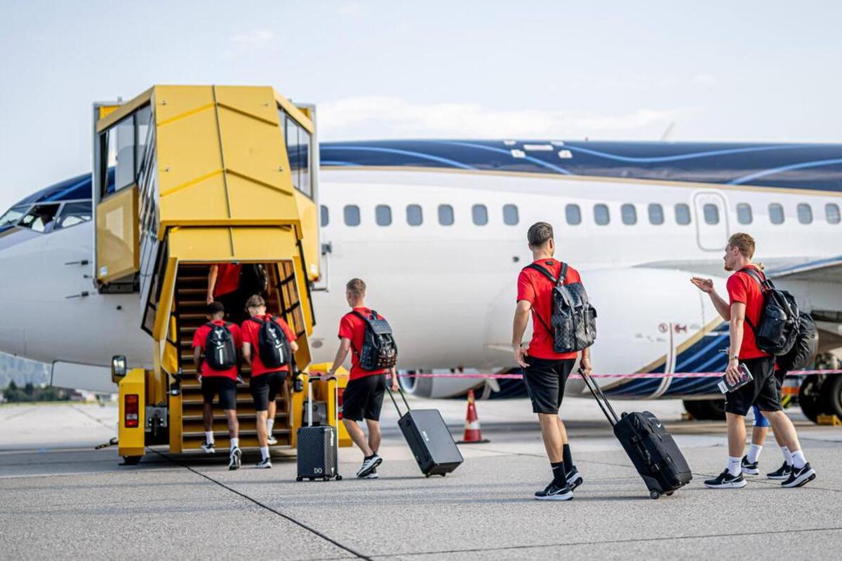 Group of young people in red shirts and black shorts boarding an airplane using a yellow mobile stairway, pulling black luggage.