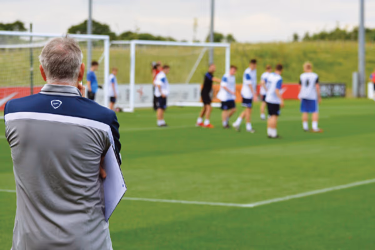 Coach observing a soccer team practicing on a green field near goalposts.