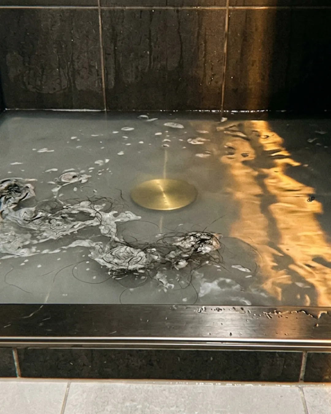 Murky water backed up around a floor drain in a dark-tiled Balmain bathroom, warm vanity light reflecting in the standing water.
