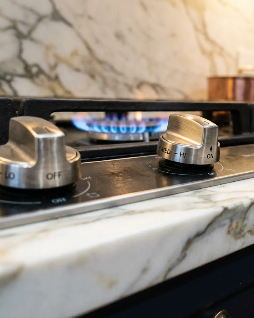Brushed stainless steel control knobs on a premium gas cooktop with a clean blue flame ring, set into marble in a Balmain terrace kitchen
