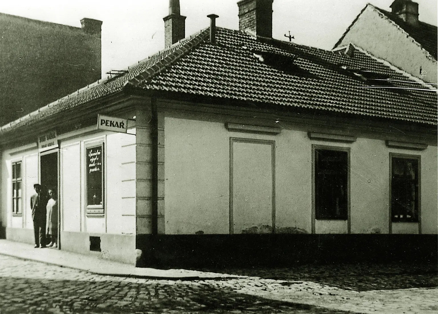 Černobílá historická fotografie rohového domu s nápisem Pekař, na verandě stojí muž a žena.
