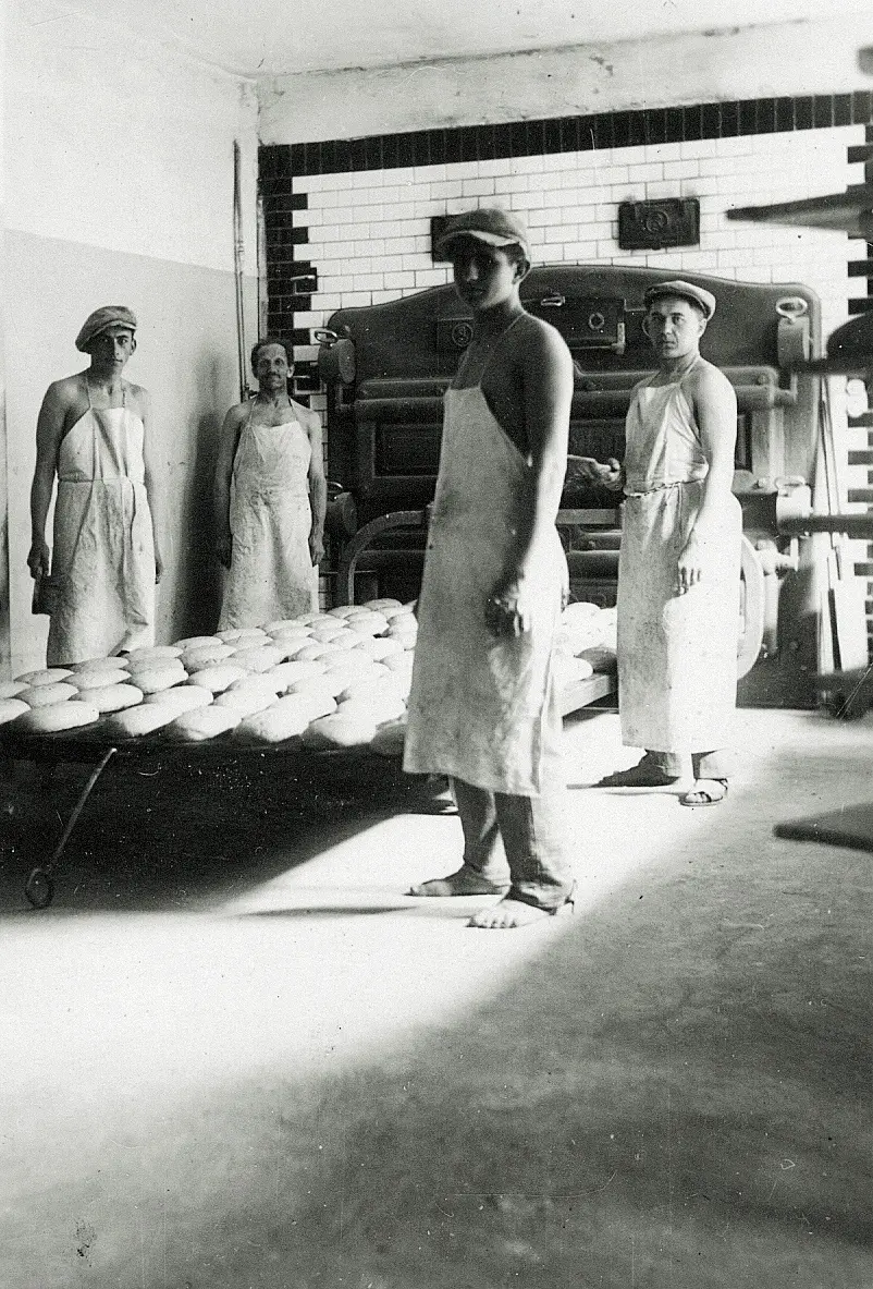 Čtyři muži v zástěrách stojí v pekárně u stolu plného bochníků chleba před velkou pecí.