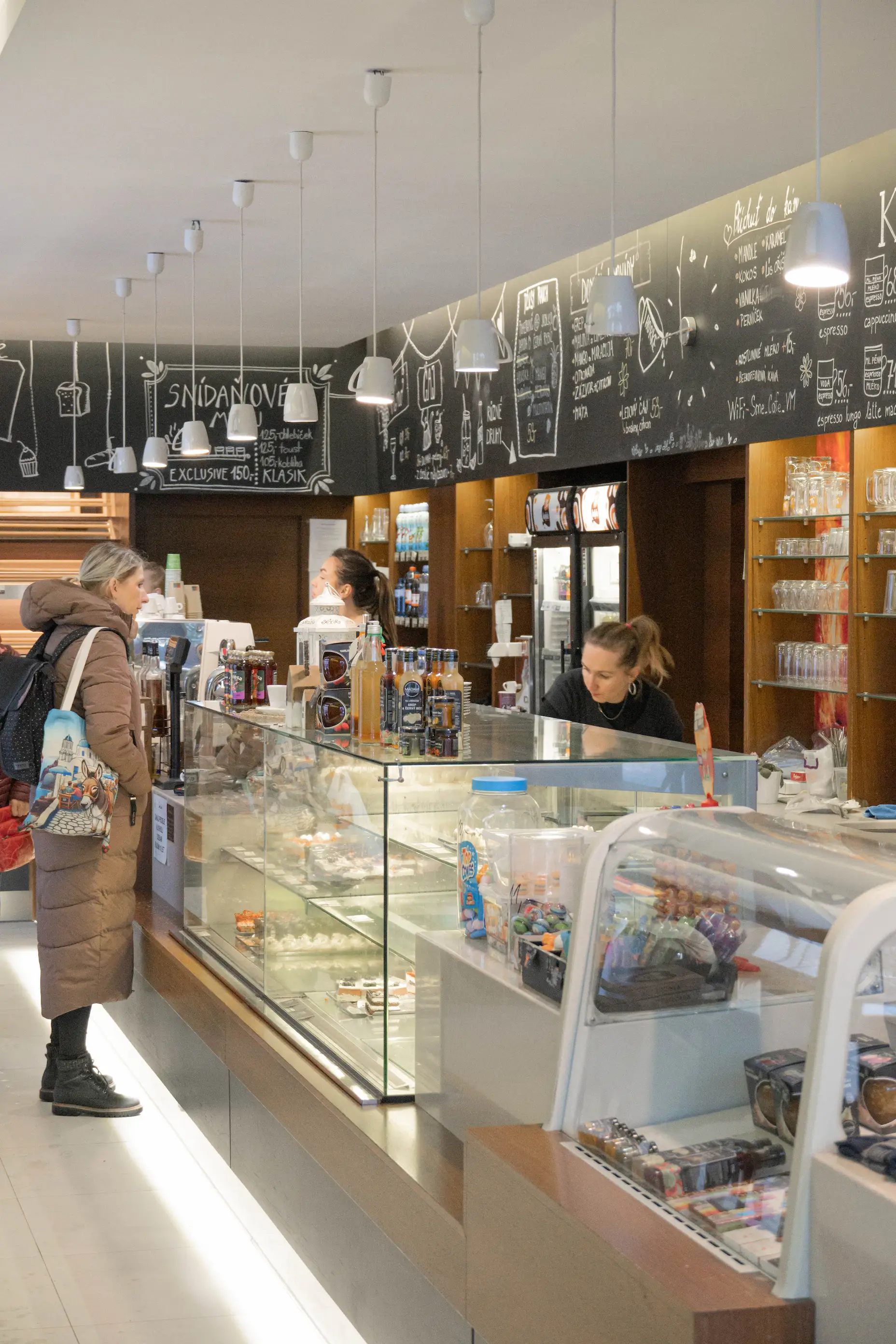 Zákaznice v zimním kabátu stojí u pultu v kavárně s nabídkou dezertů a nápojů, dvě servírky obsluhují za pultem.