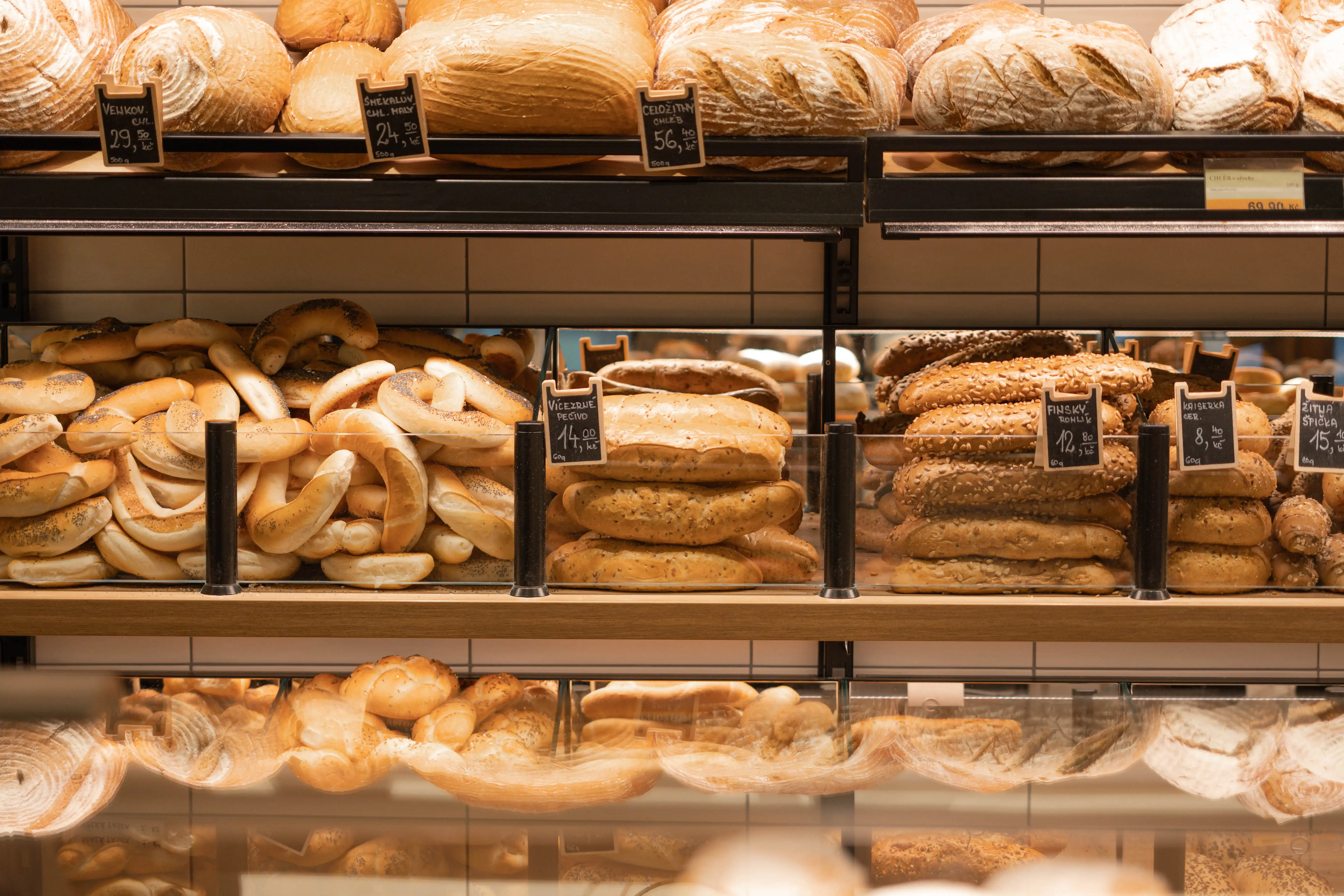 Různé druhy čerstvého pečiva v regálech pekárny s cenovkami v korunách.