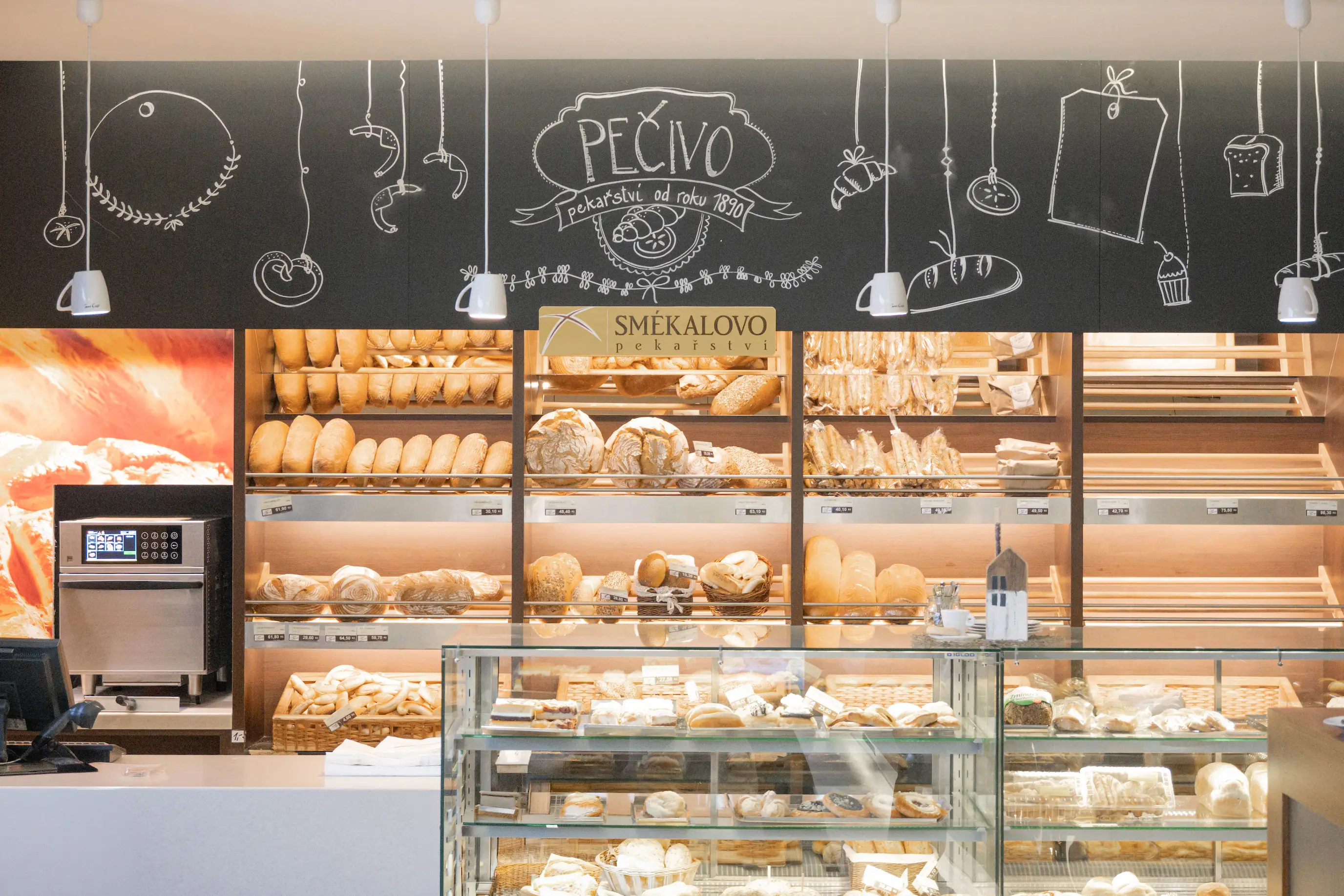 Pekárna s pultem plným různých druhů pečiva a chlebů, s kresbami pečiva na černé zdi za pultem.