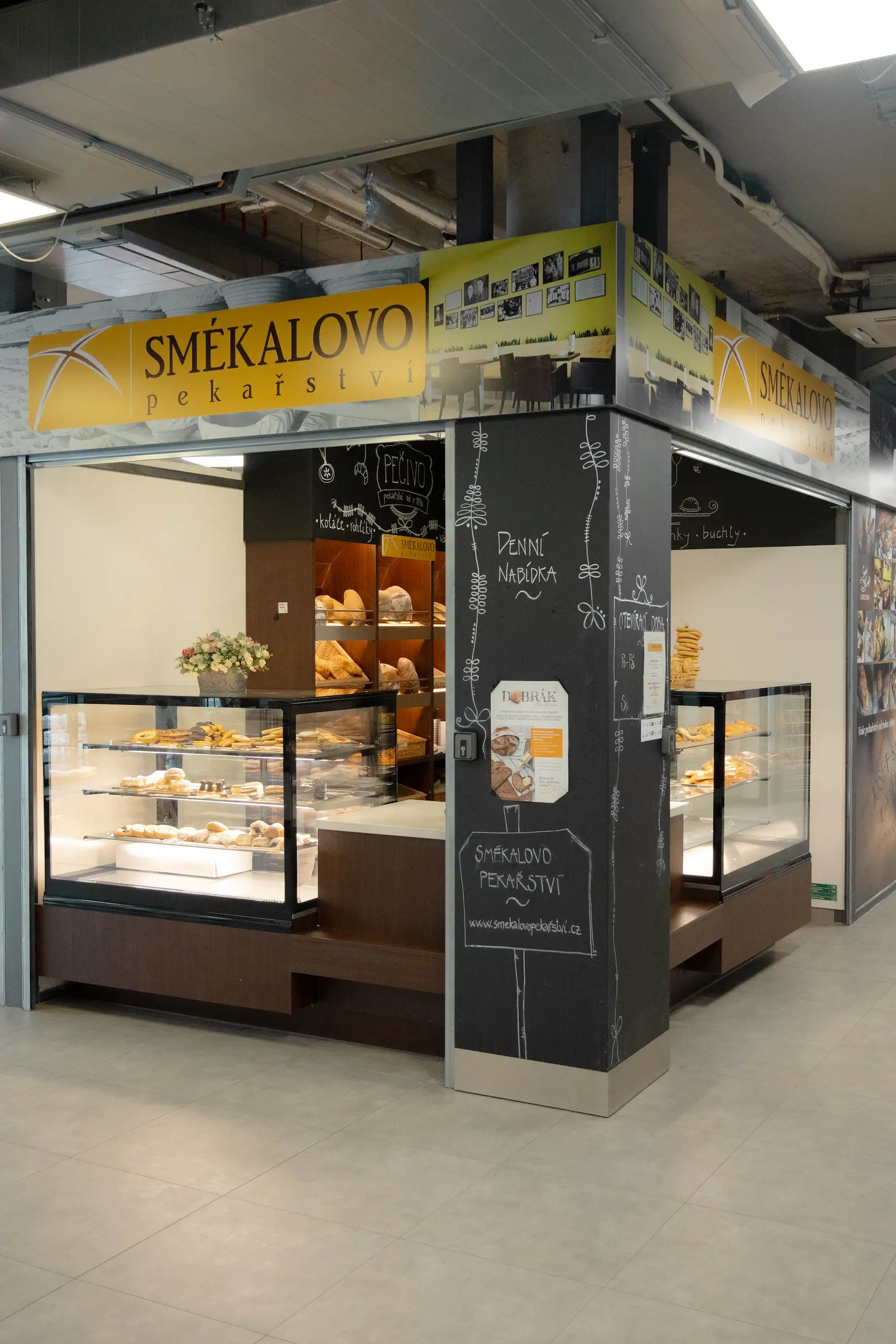 Městský stánek Smékalovo pekařství s vitrínami plnými pečiva a sladkostí, černá stěna s nápisy denní nabídky.