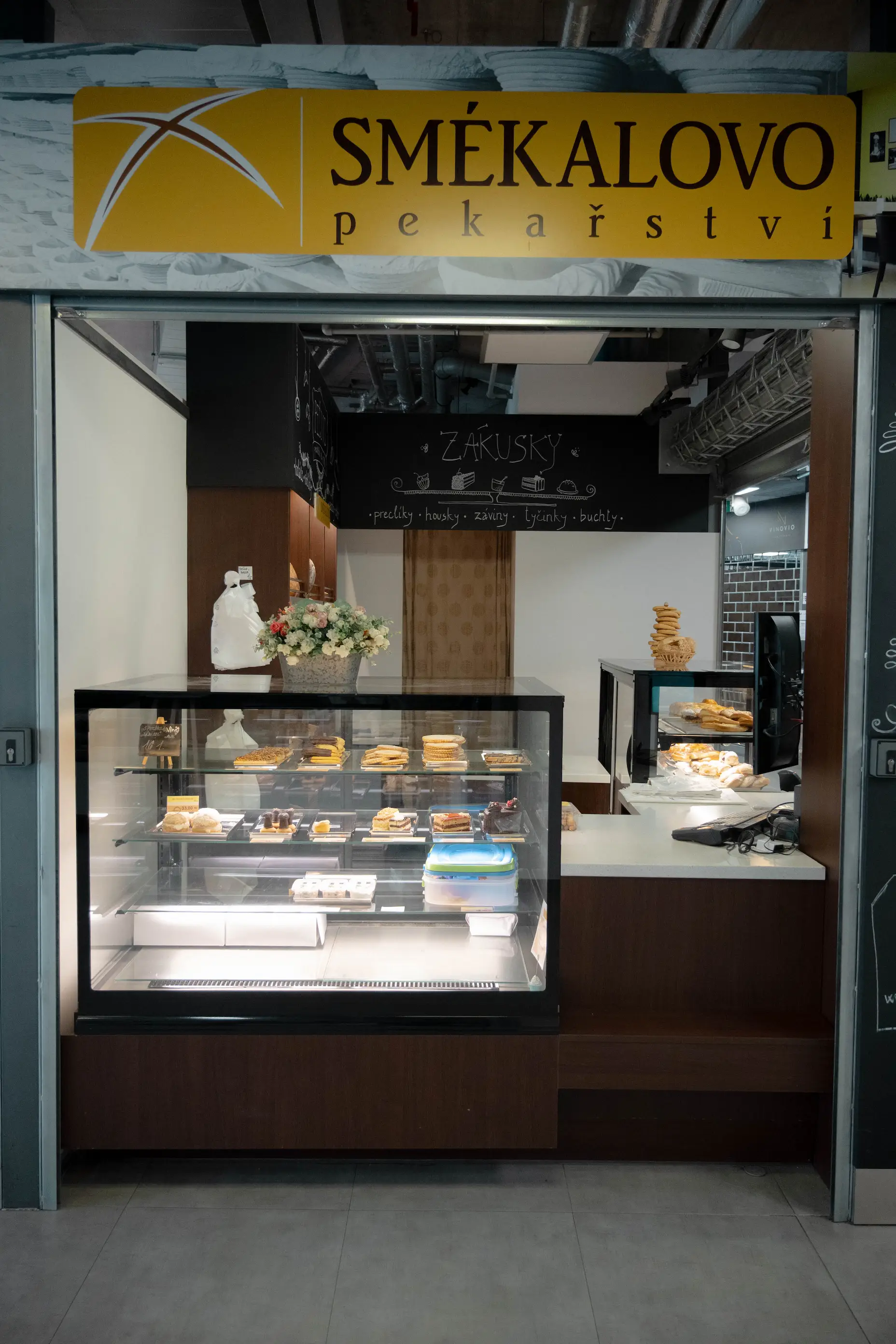 Pekárna Smékalovo s vitrínou plnou pečiva a dortíků, vedle květinová dekorace.