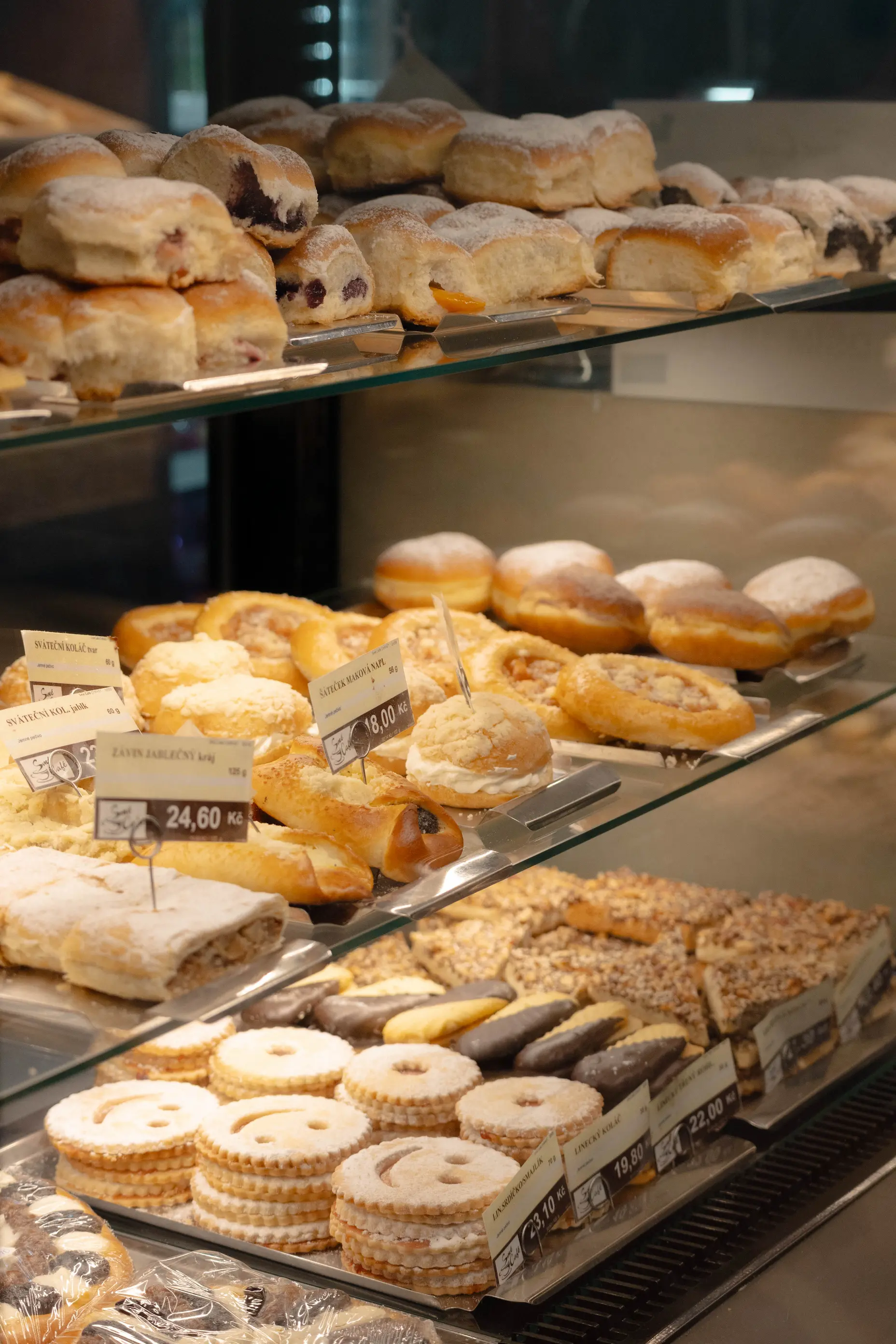 Skleněná vitrína v pekárně plná různých druhů českého pečiva a sladkostí, včetně posypaných buchet, koláčů a sušenek s cenovkami v korunách.