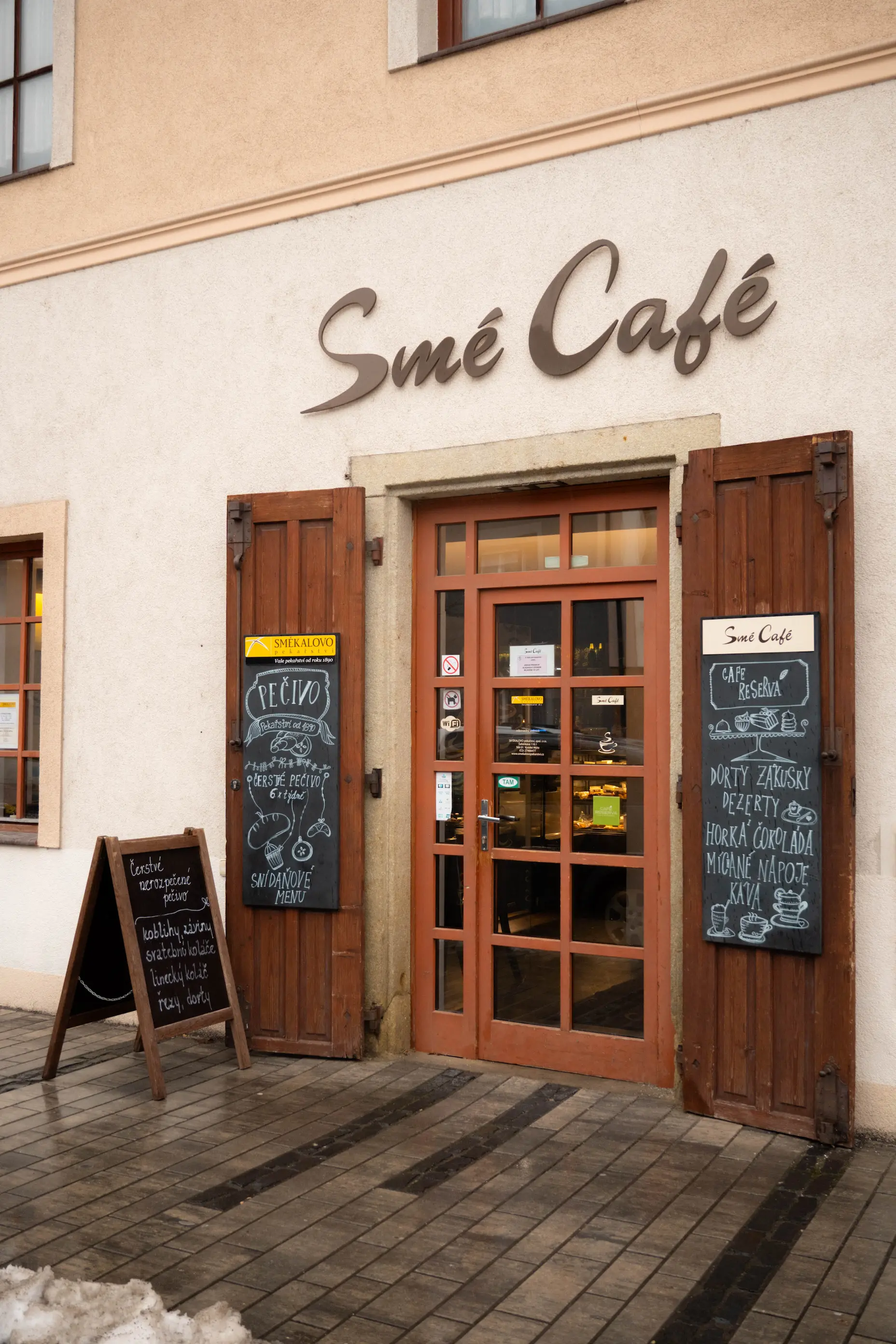 Vstup do kavárny Smé Café s dřevěnými dveřmi a dvěma tabulemi s nabídkou pečiva, dortů a nápojů.