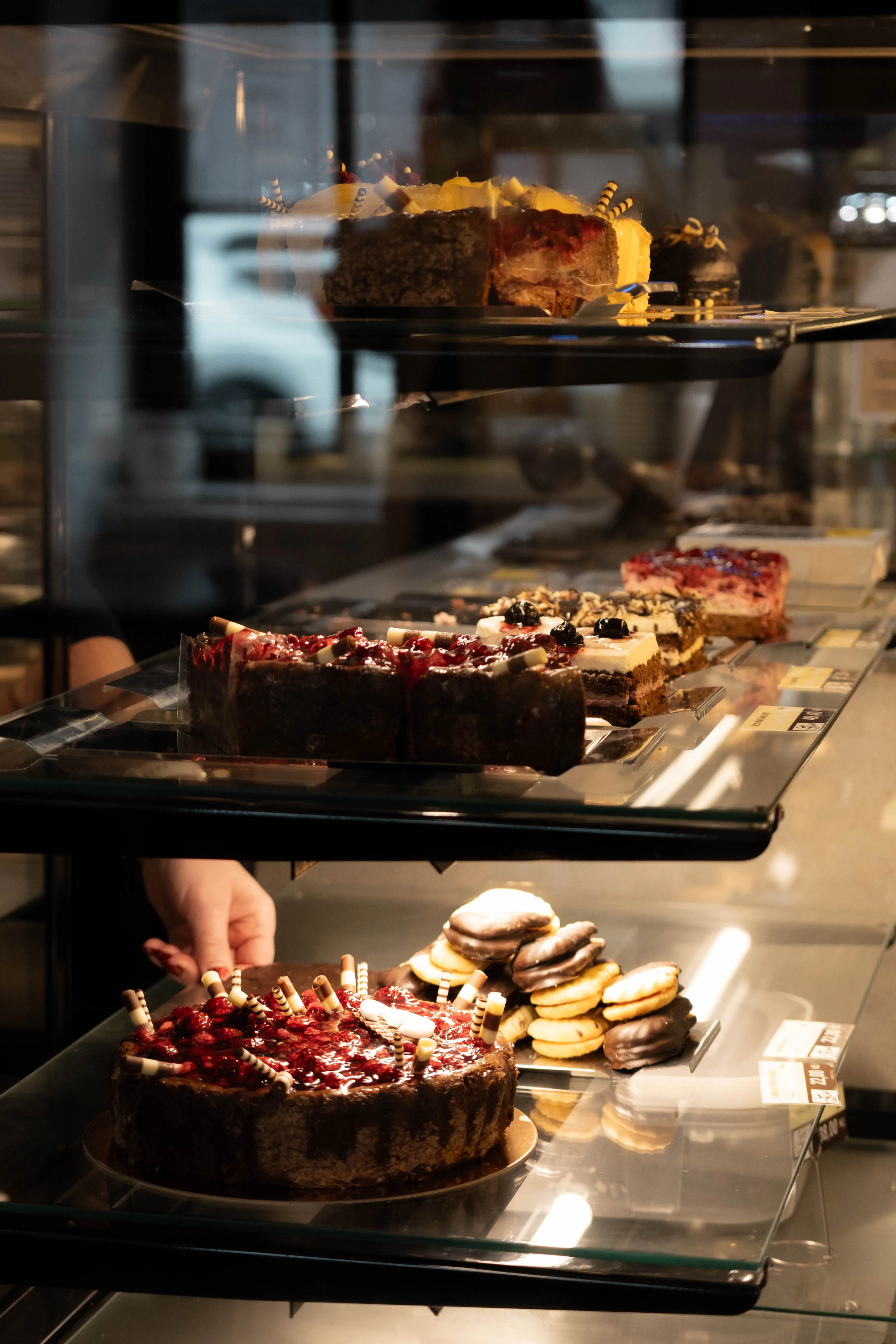 Skleněná vitrína v cukrárně plná různých dortů a sušenek s čokoládovou polevou, za skllem vidět ruku prodavače.