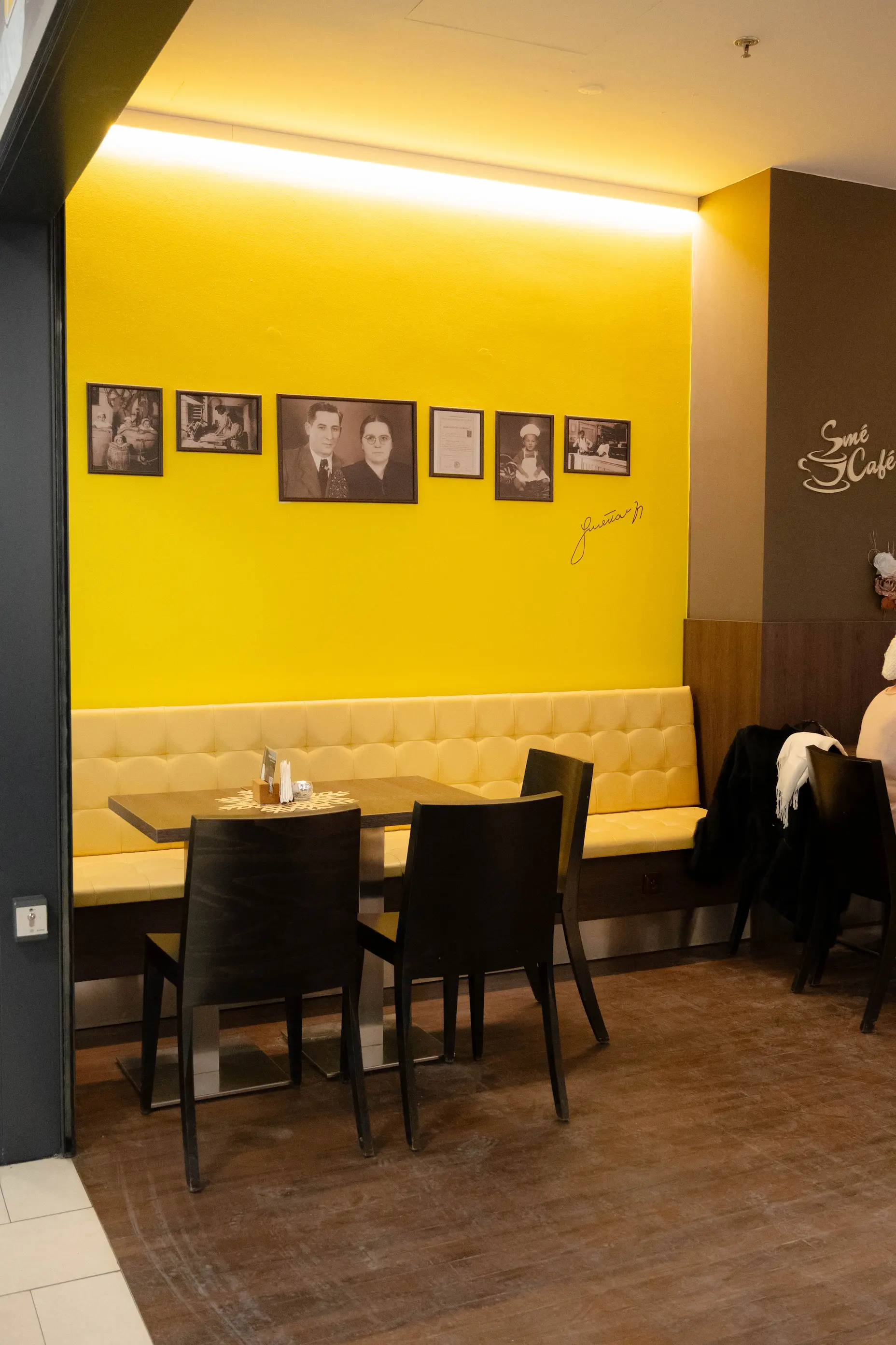 Interiér kavárny s žlutou stěnou, na které visí černobílé fotografie a žlutou lavicí s tmavými židlemi u dřevěného stolu.