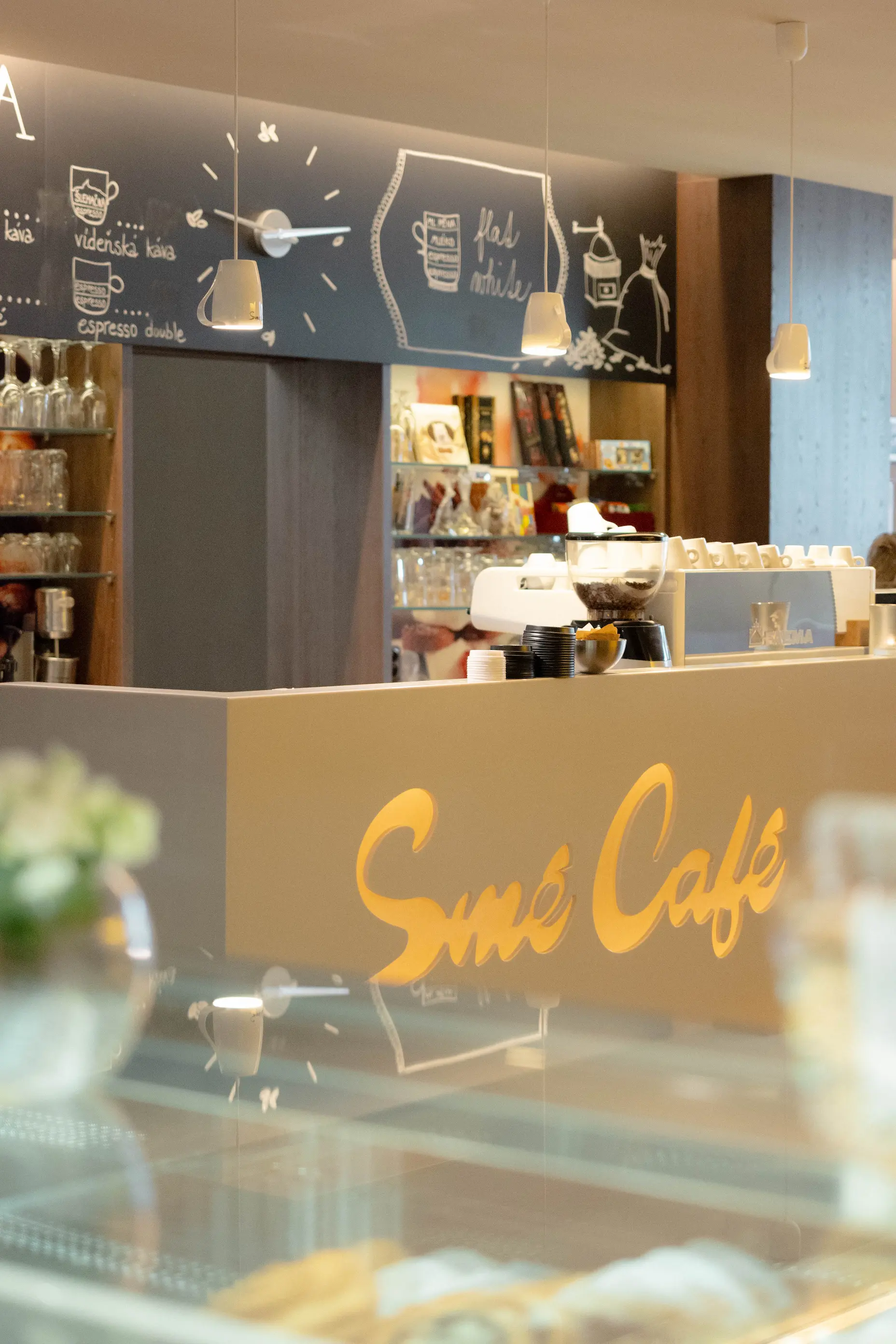 Interiér kavárny s pultem a nápisem Smé Café, visící světla a černá tabule s nabídkou kávy na pozadí.