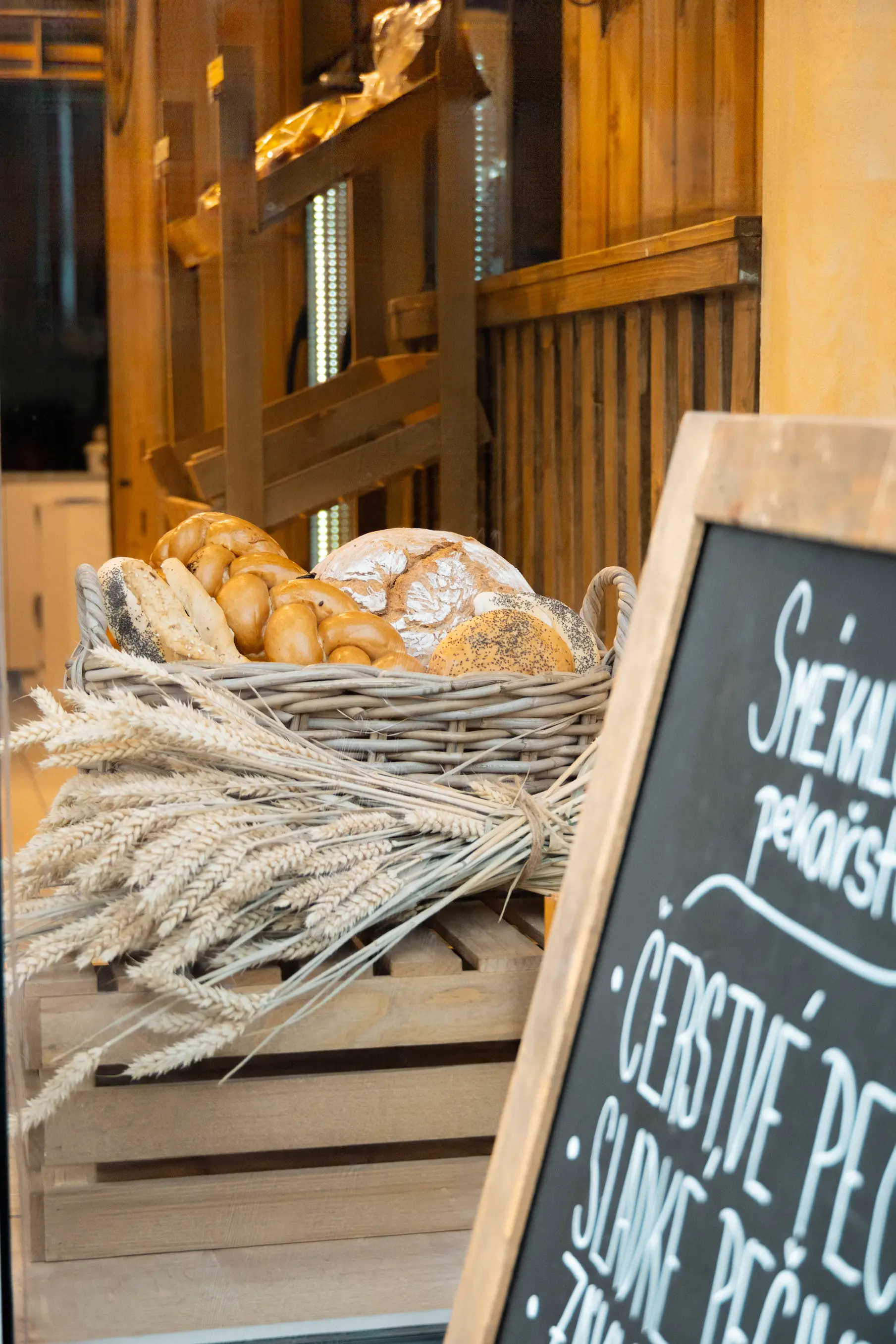 Koš plný různých druhů pečiva vedle náznaku tabule s českým textem v pekárně.