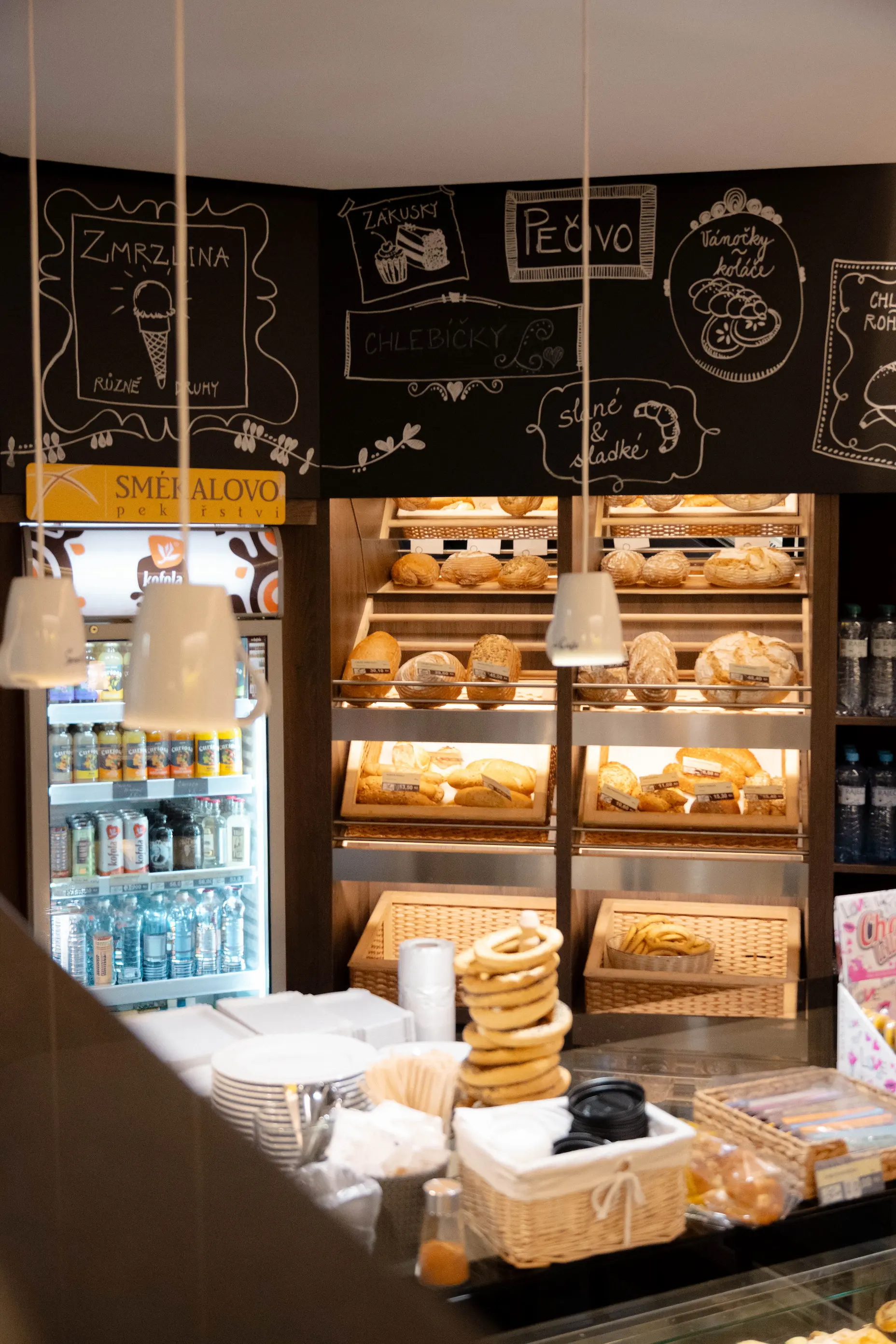 Pekařství s regály plnými různých druhů pečiva a nápoji ve výloze, s dekoracemi a cedulemi na zdi v pozadí.