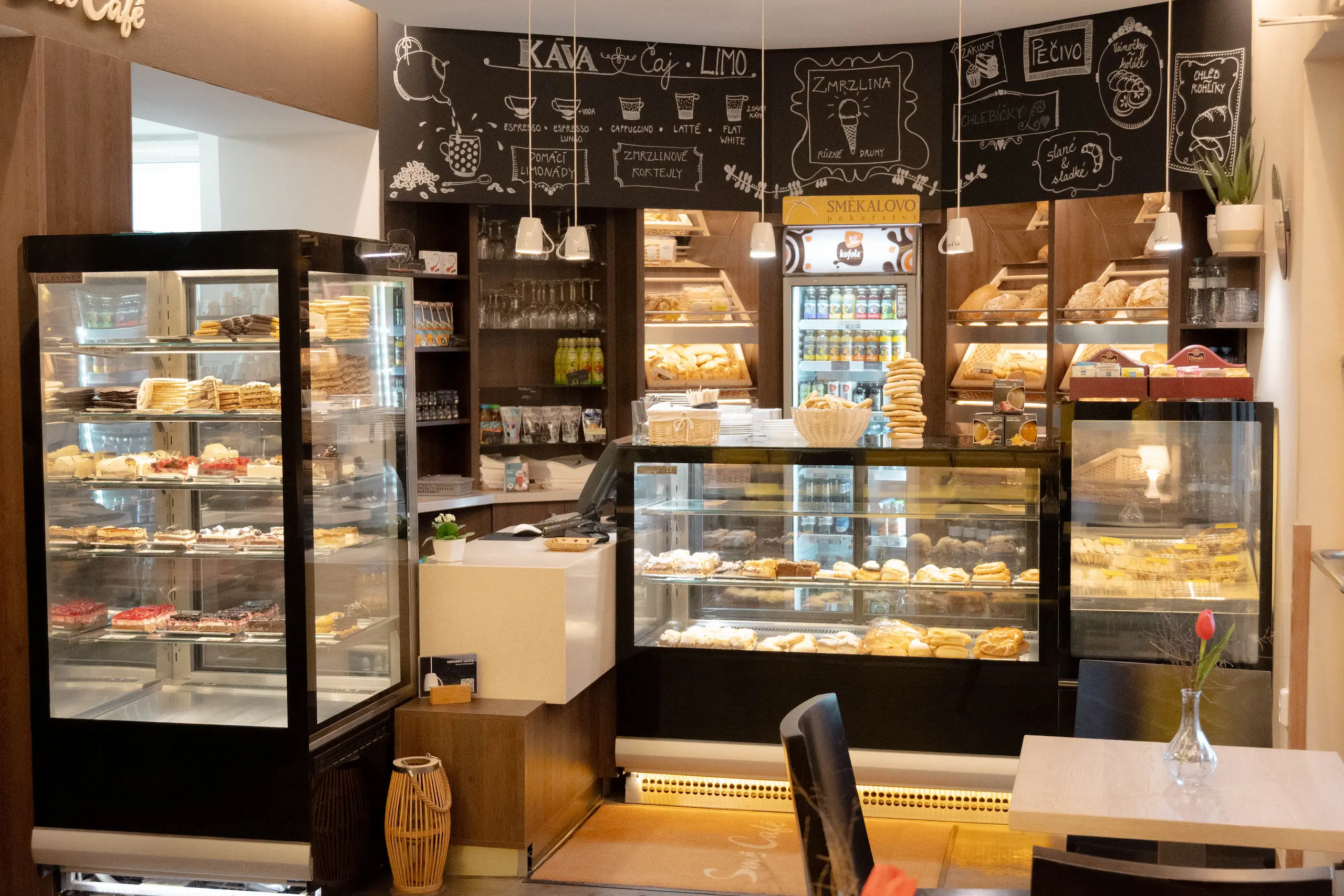 Interiér kavárny s vystavenými zákusky a pečivem ve vitrínách, s tabulovou nabídkou nápojů a jídel na pozadí.