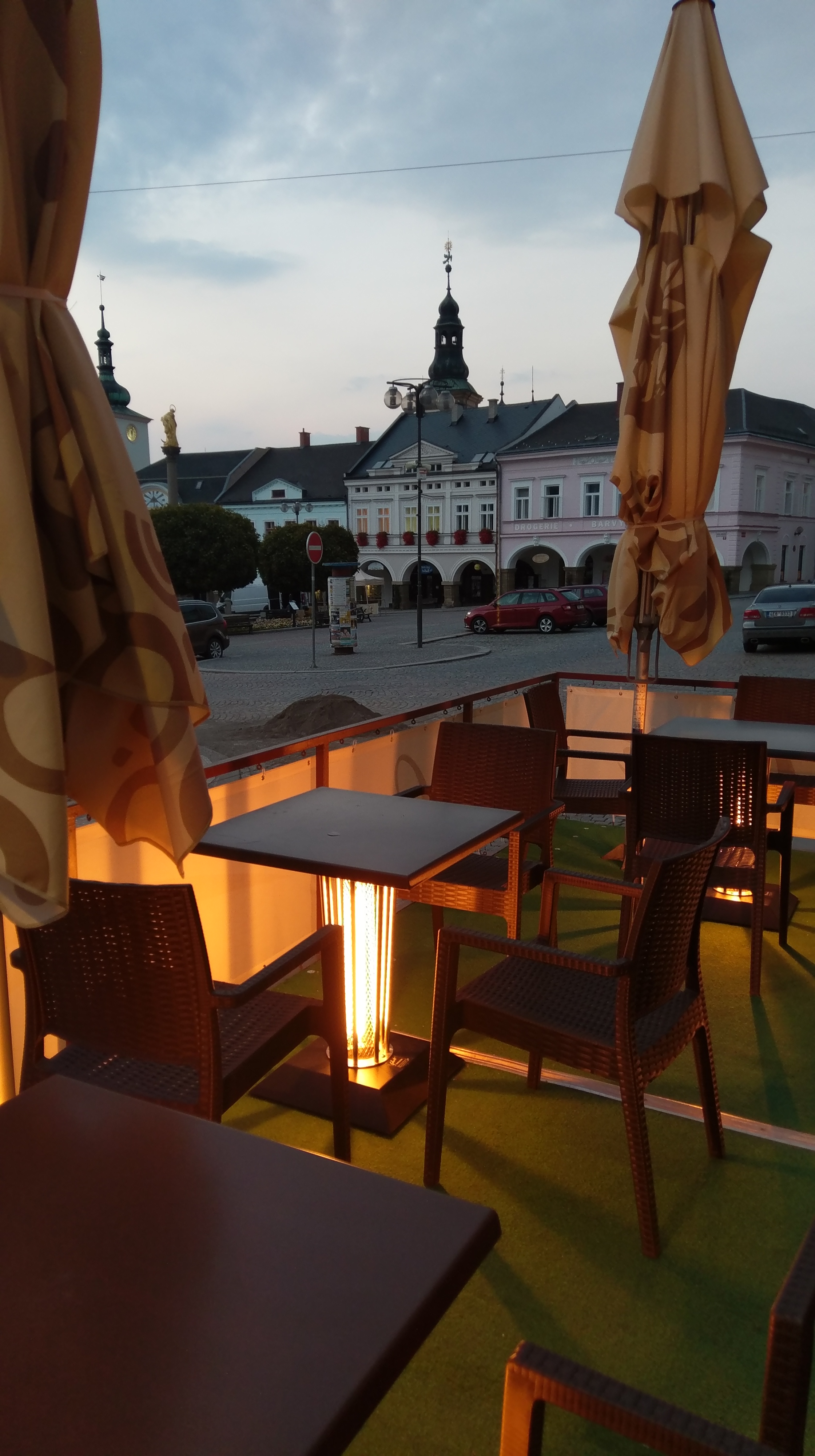 Venkovní posezení kavárny s prázdnými židlemi a stolky, podvečerní pohled na historické budovy na náměstí v pozadí.