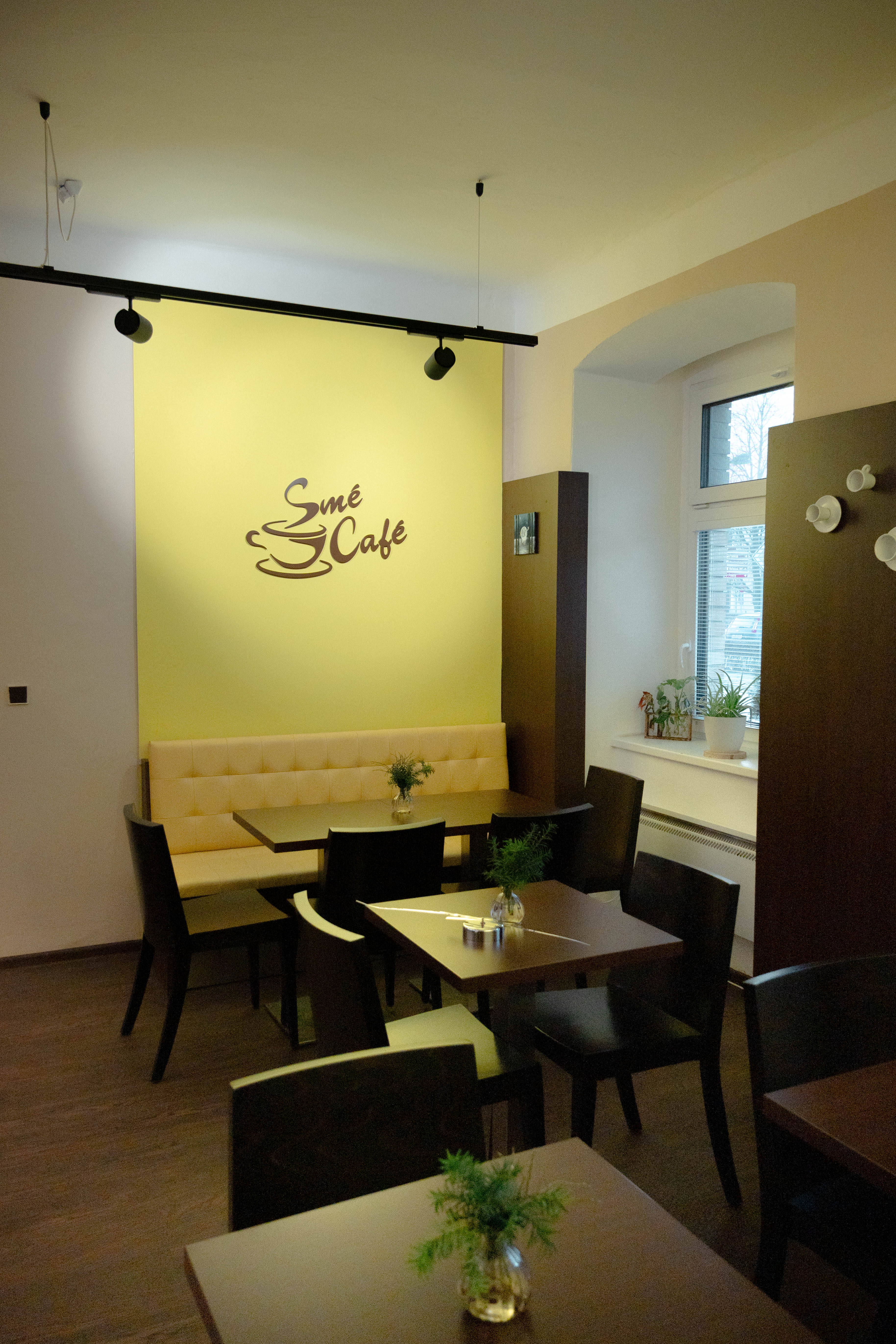 Interiér kavárny s tmavým nábytkem a žlutou stěnou s nápisem Smé Café.