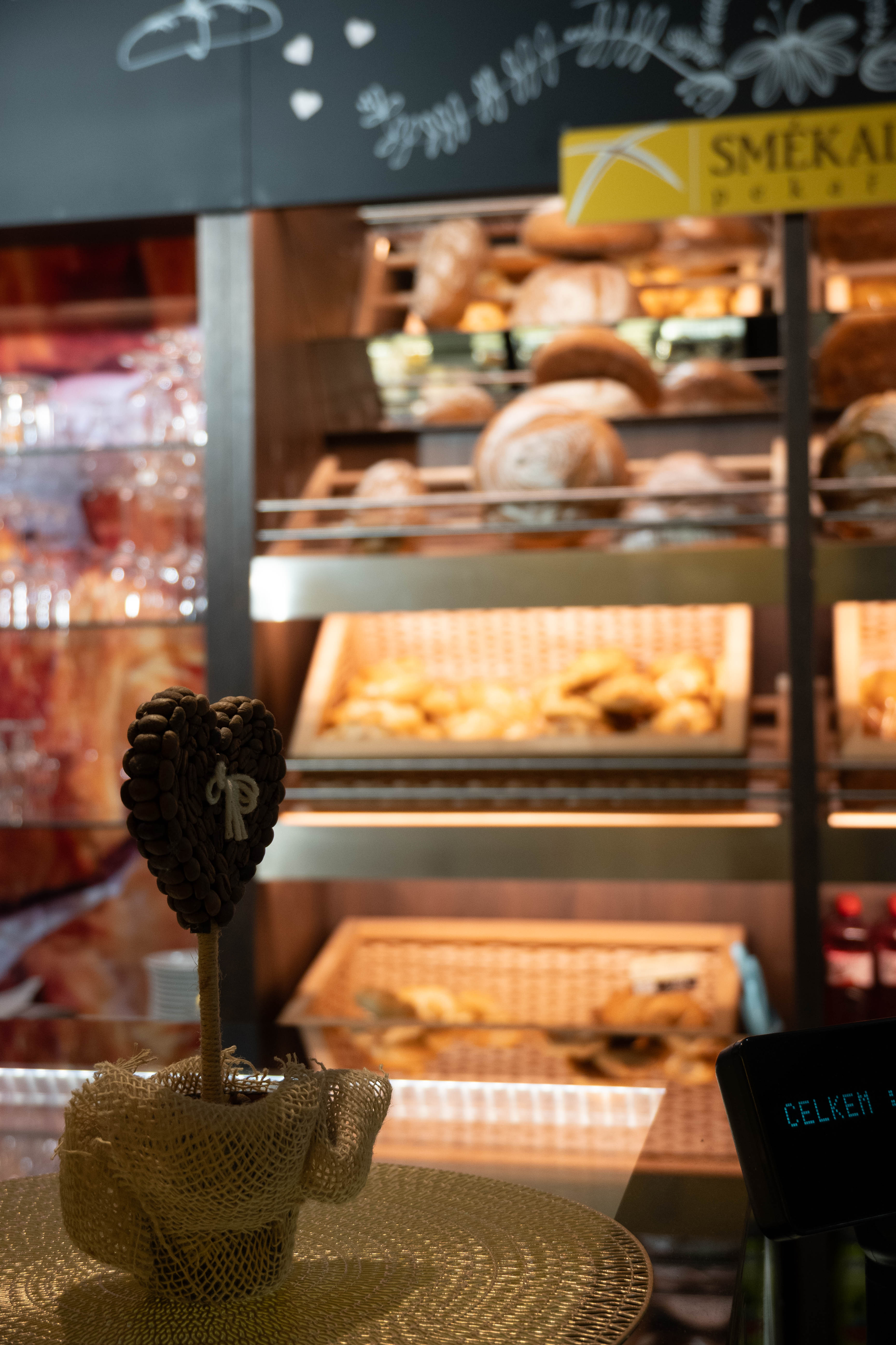 Dekorace srdce z kávových zrn v síťovaném obalu na stole v kavárně, v pozadí police s pečivem.