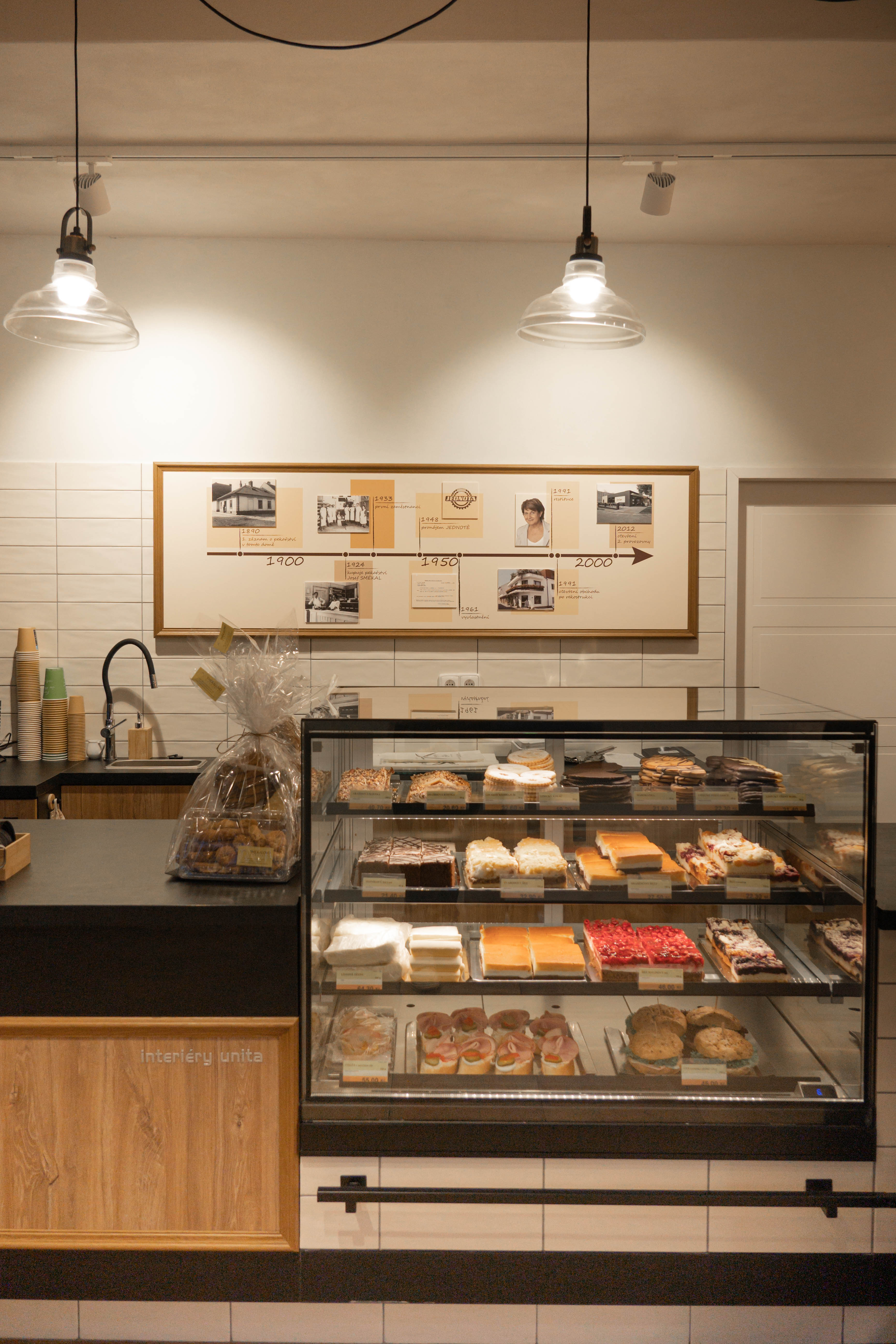 Pekařská prodejna s vitrínou plnou různých pečiv a sladkých dezertů, nad pultem je nástěnka s časovou osou historie.