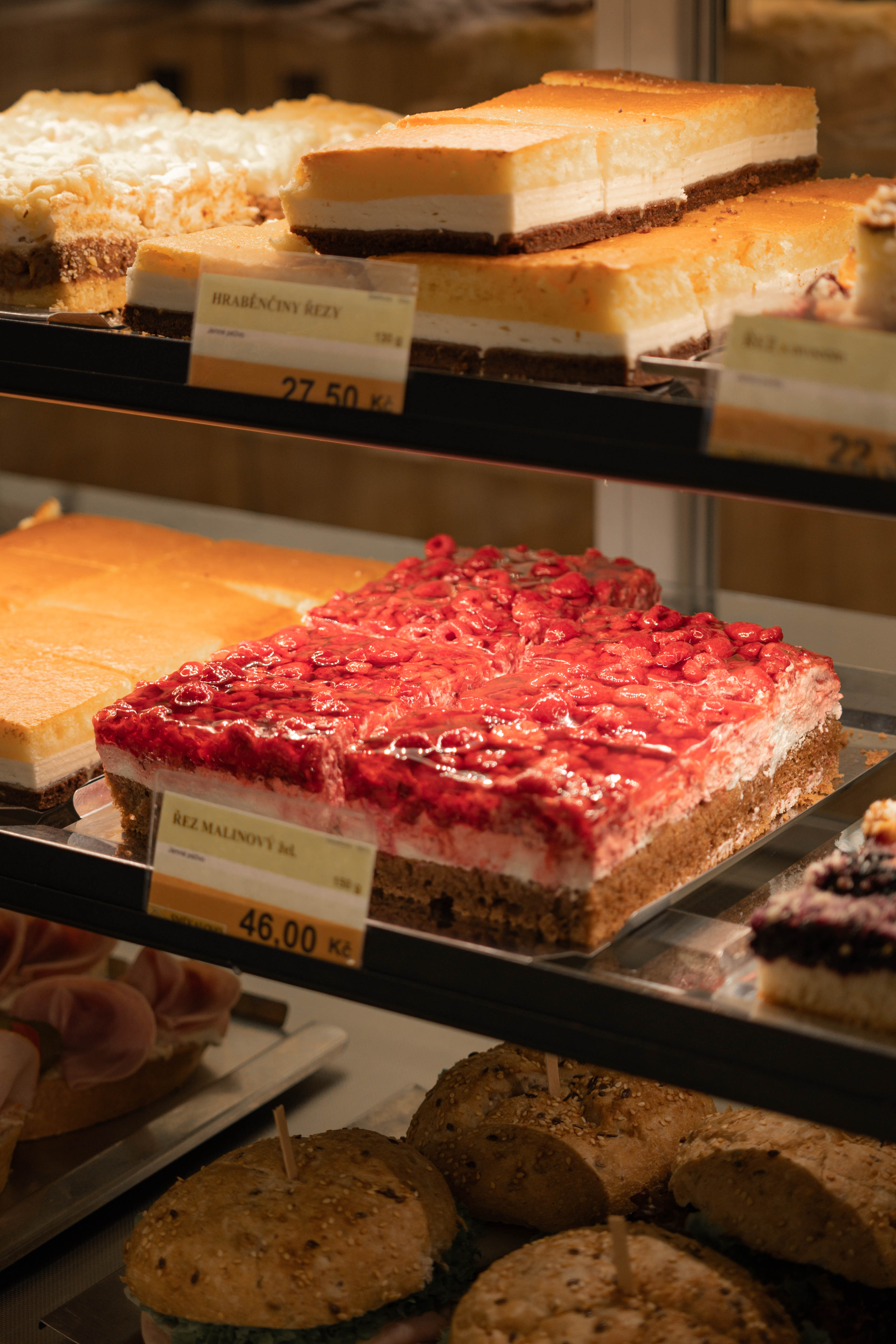 Různé druhy dortů, včetně malinového řezu s lesními plody, vystavené v cukrárenské vitríně.