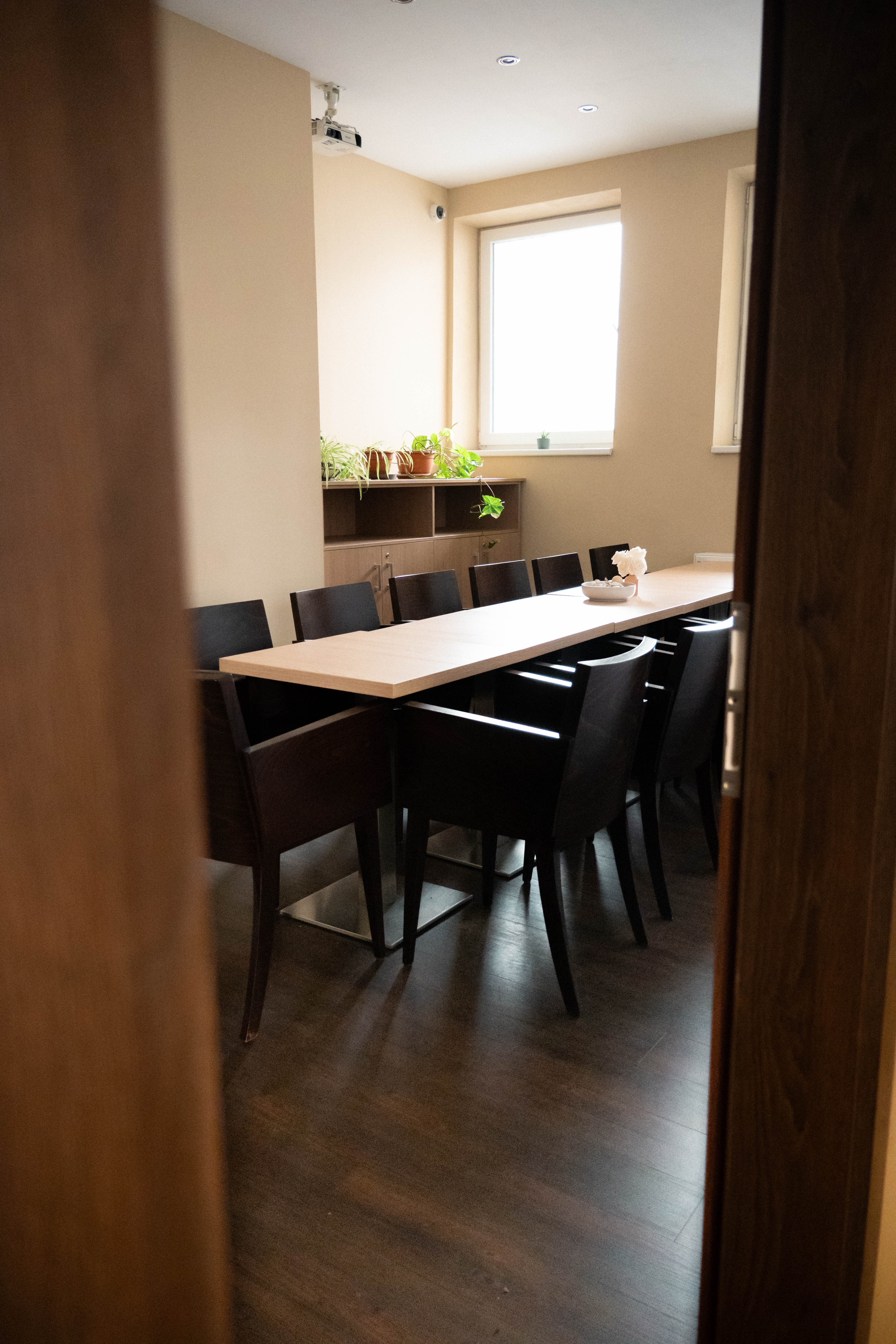 Moderní zasedací místnost s dlouhým světlým stolem a tmavými židlemi, na stole dekorace a za oknem police s květinami.