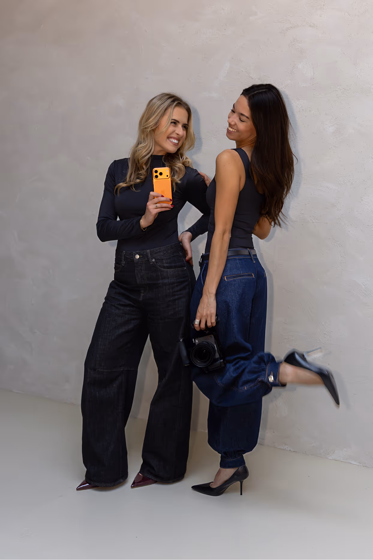 Two women posing smiling; one holds an orange smartphone and the other holds a camera, both wearing black tops and dark wide-leg jeans.