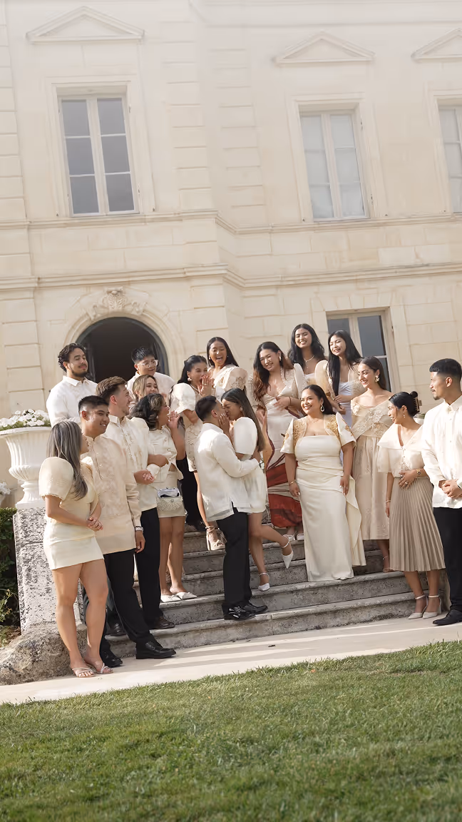 A group of people dressed in traditional Filipino attire, with the couple kissing in the center on stone steps in front of a classical building.