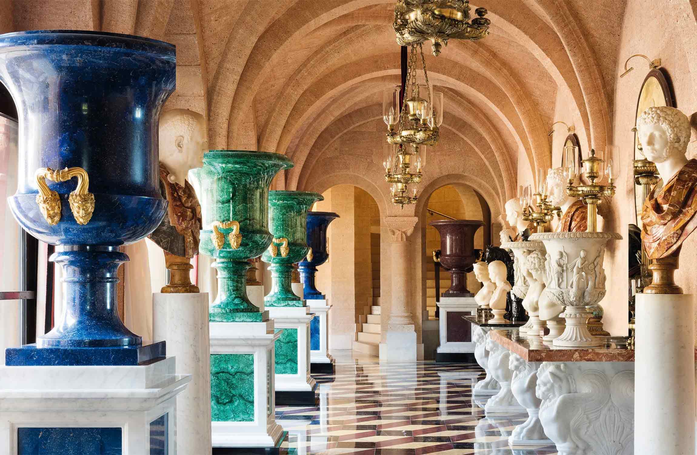 Luxury Renaissance-style gallery featuring monumental marble vases, vaulted arches, classical sculptures, and polished geometric flooring inside an aristocratic Mediterranean estate.
