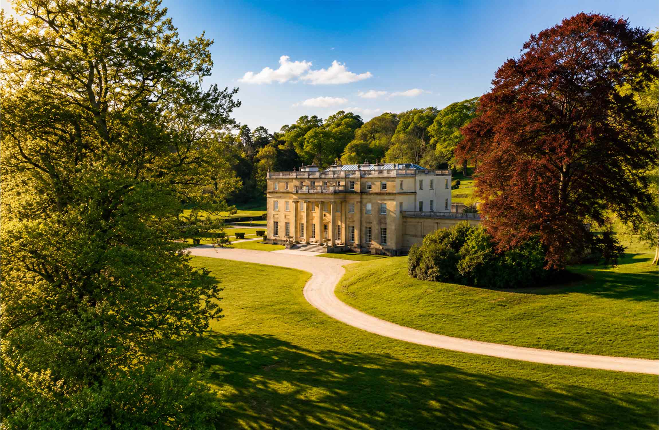 Aerial view of a $70 million luxury manor in the Cotswolds, surrounded by rolling green countryside, mature trees, and elegant landscaped gardens in England.