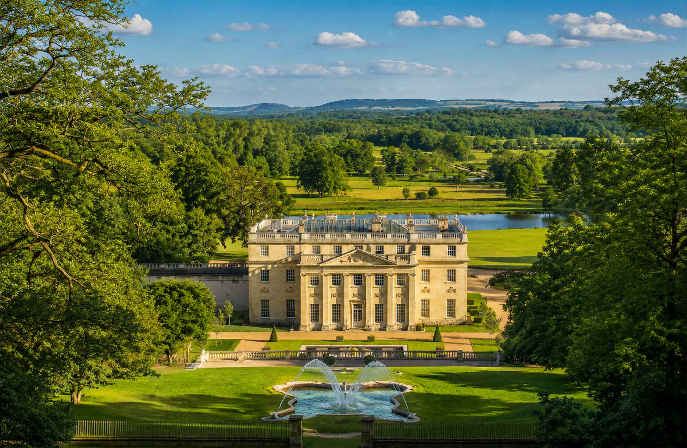 Panoramic aerial view of a $70 million luxury manor estate in the Cotswolds, featuring formal gardens, elegant fountains, a private lake, and the rolling English countryside.