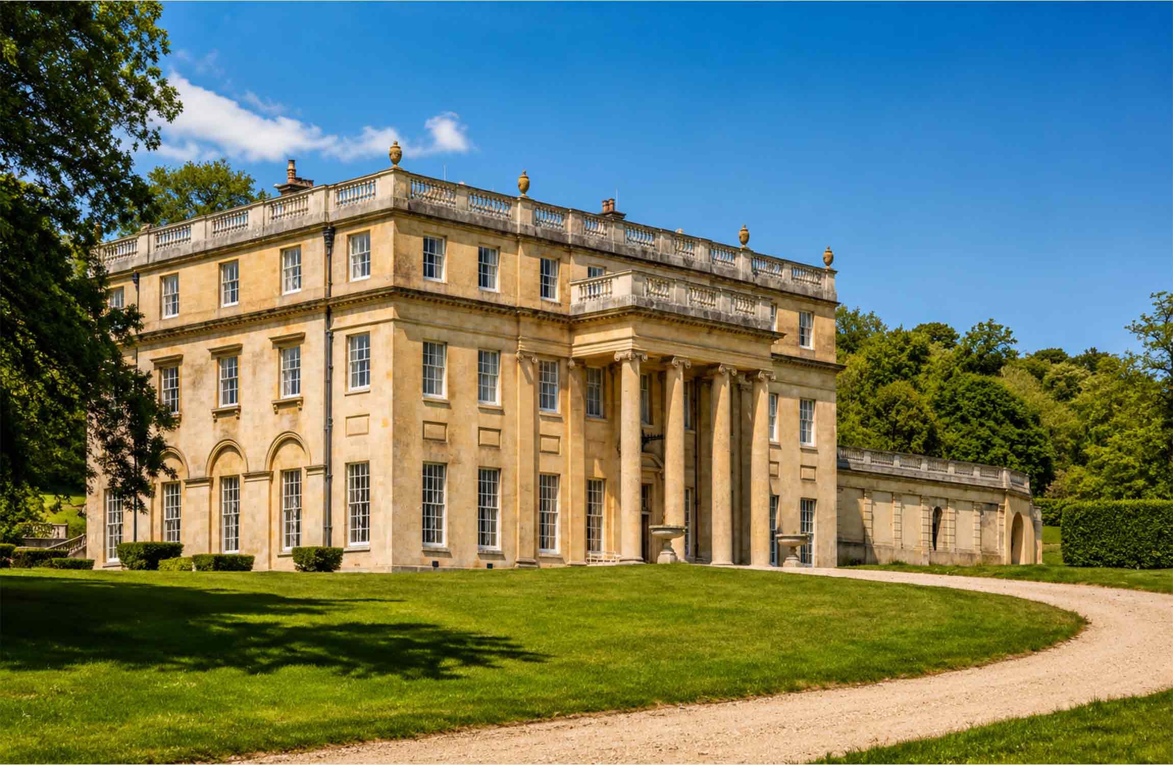 Front view of a $70 million neoclassical manor in the Cotswolds featuring elegant stone architecture, grand columns, landscaped gardens, and a private driveway in the English countryside.