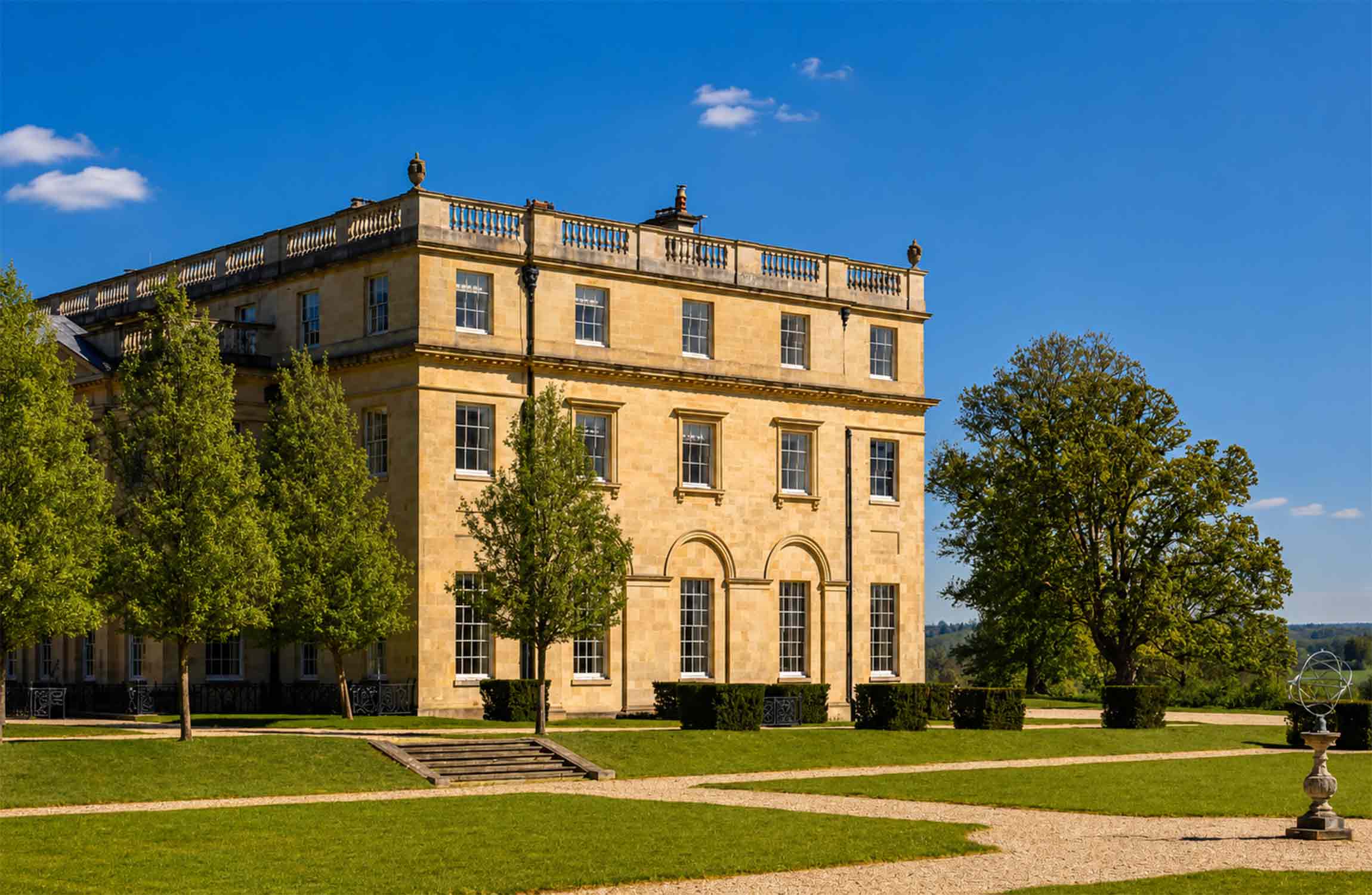 Side view of a $70 million luxury manor in the Cotswolds showcasing elegant neoclassical architecture, landscaped gardens, mature trees, and the peaceful English countryside.