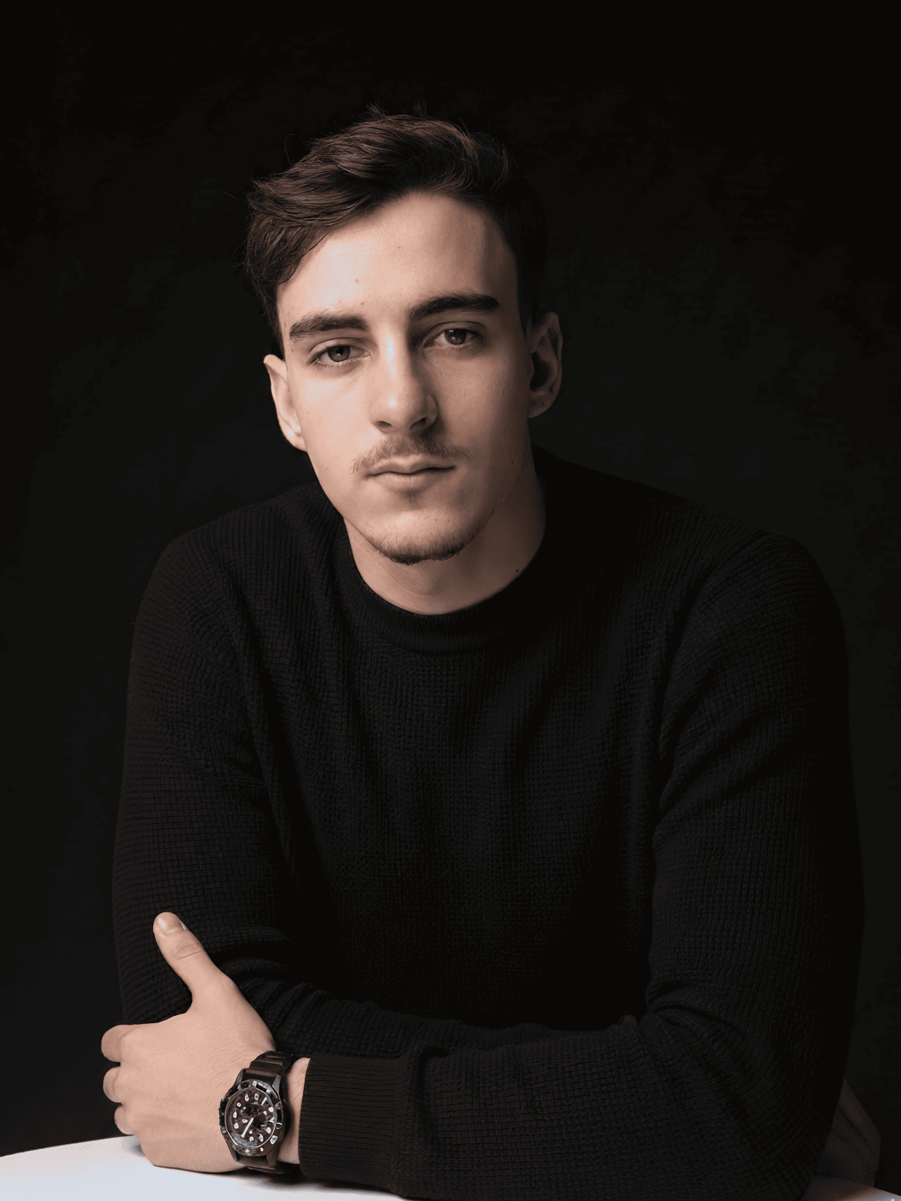 Young man with short brown hair wearing a black sweater and a wristwatch, posing against a black background.