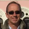 Middle-aged man wearing sunglasses and a dark jacket, smiling outdoors in front of a building with arched windows.