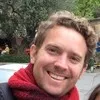 Smiling man with curly hair wearing a maroon scarf outdoors with greenery in the background.
