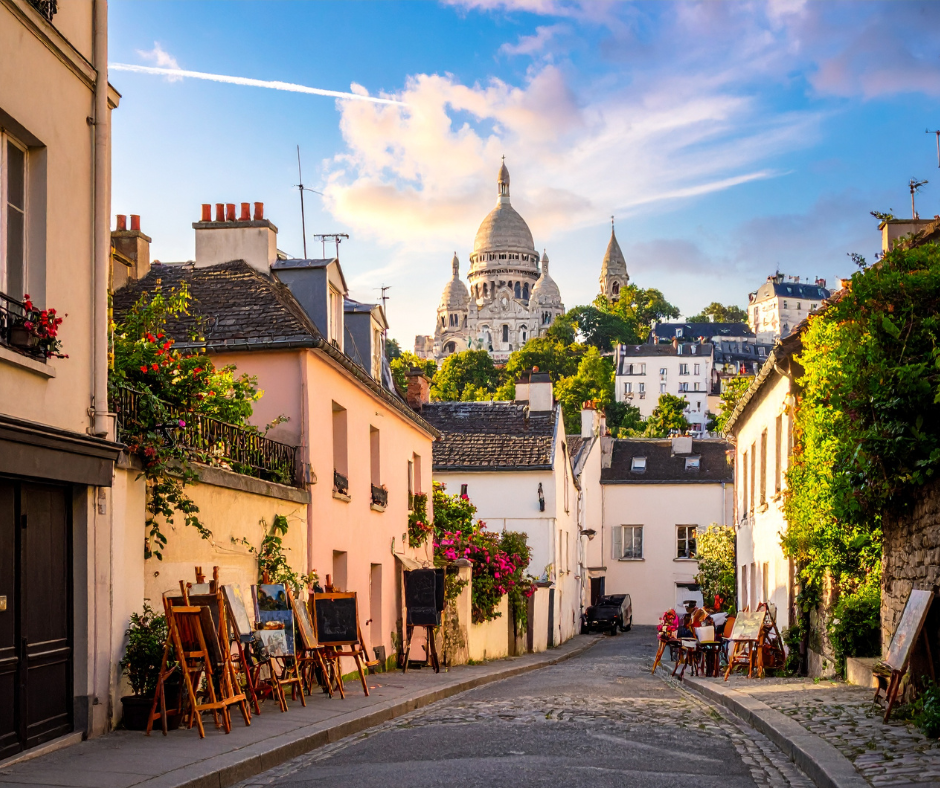 Picturesque street in Montmartre, Paris with easels displaying artwork on the sidewalk and the Sacré-Cœur Basilica visible in the background under a partly cloudy sky.