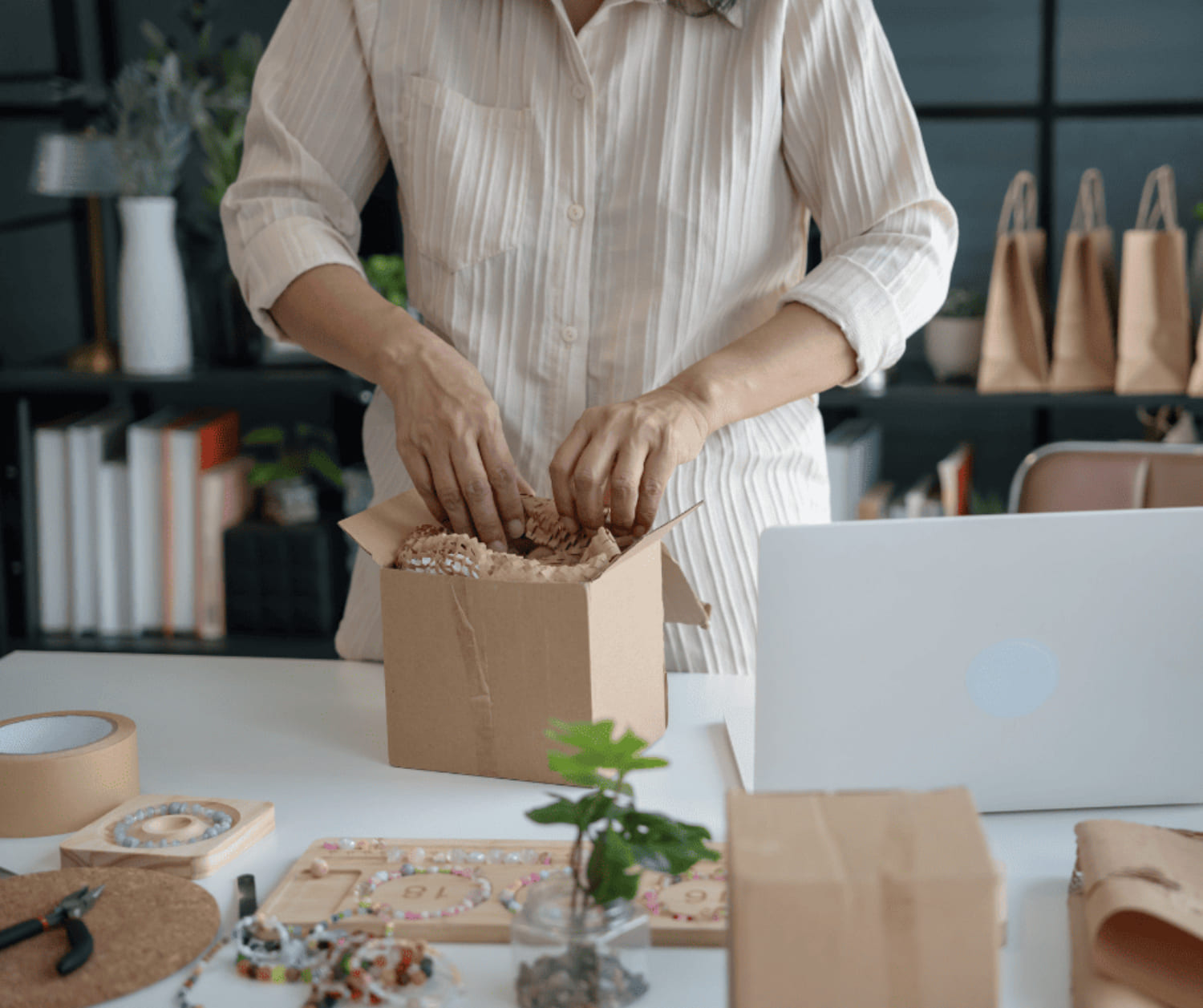 Person packing a cardboard box with protective paper on a table with beads, tools, and a laptop.