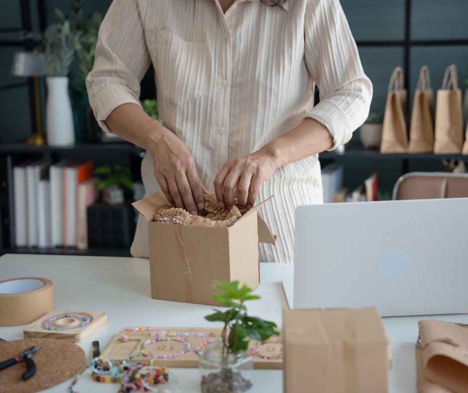 Person packing a cardboard box with crinkle paper padding on a table surrounded by crafting materials and a laptop.