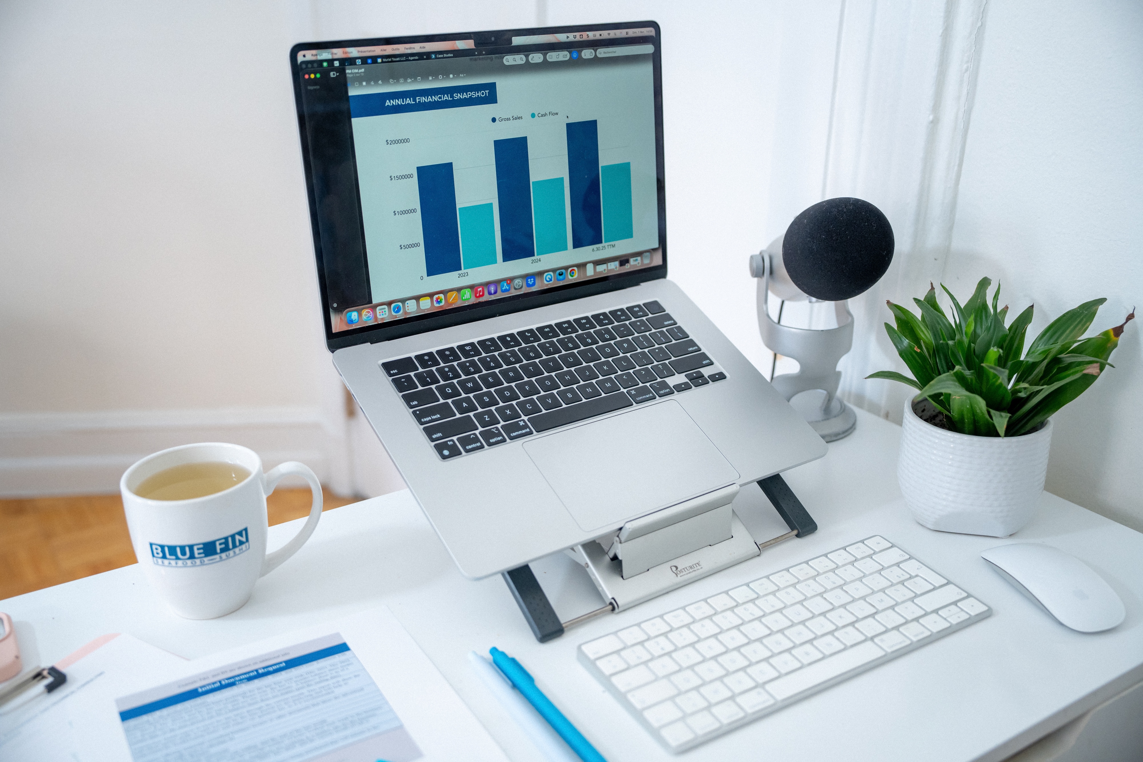 Laptop on a white desk displaying an annual financial snapshot bar chart, next to a microphone, potted plant, keyboard, mouse, and a Blue Fin Seafood Supply coffee mug.