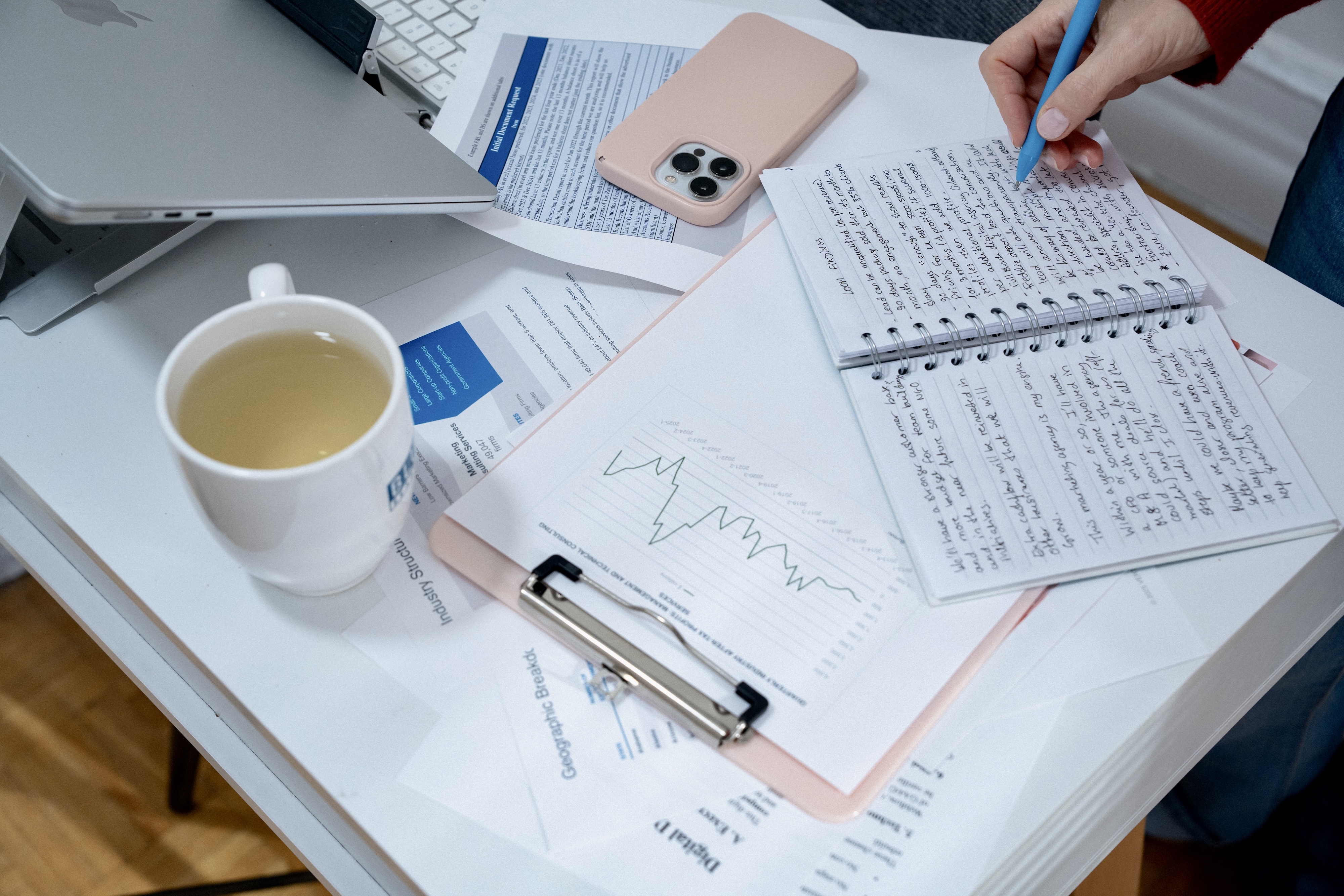 Person writing notes in a spiral notebook on a desk with a tablet, smartphone, chart papers, and a cup of tea.