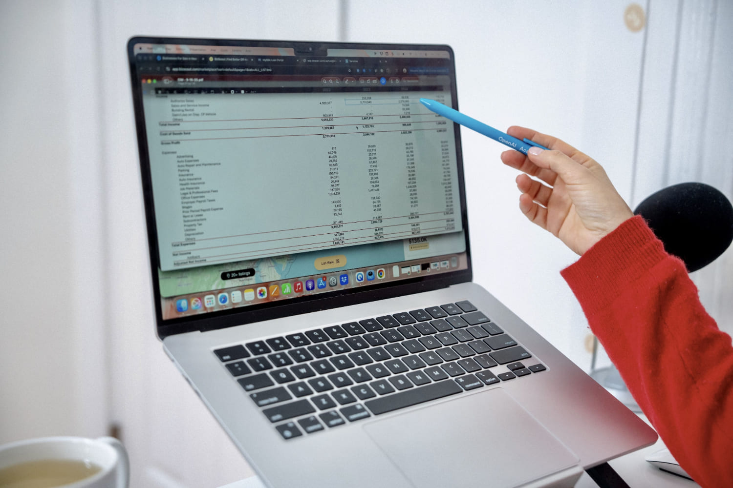 Hand in red sweater holding a blue OpenAI pen pointing at financial data on a laptop screen.