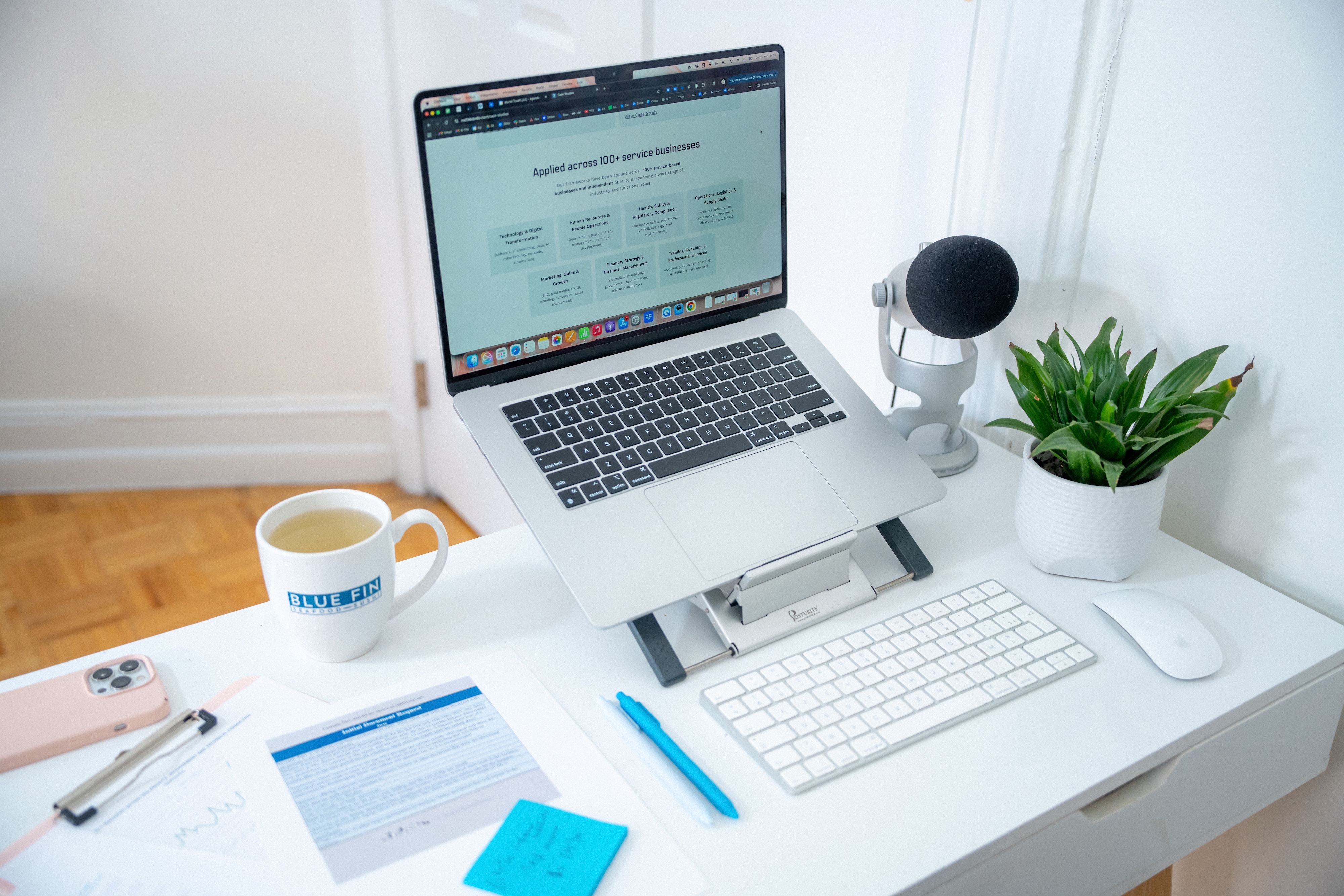 Modern white desk with a laptop on a stand, wireless keyboard and mouse, a microphone, potted plant, clipboard with papers, blue pen, sticky notes, smartphone, and a mug labeled BLUE FIN filled with tea.