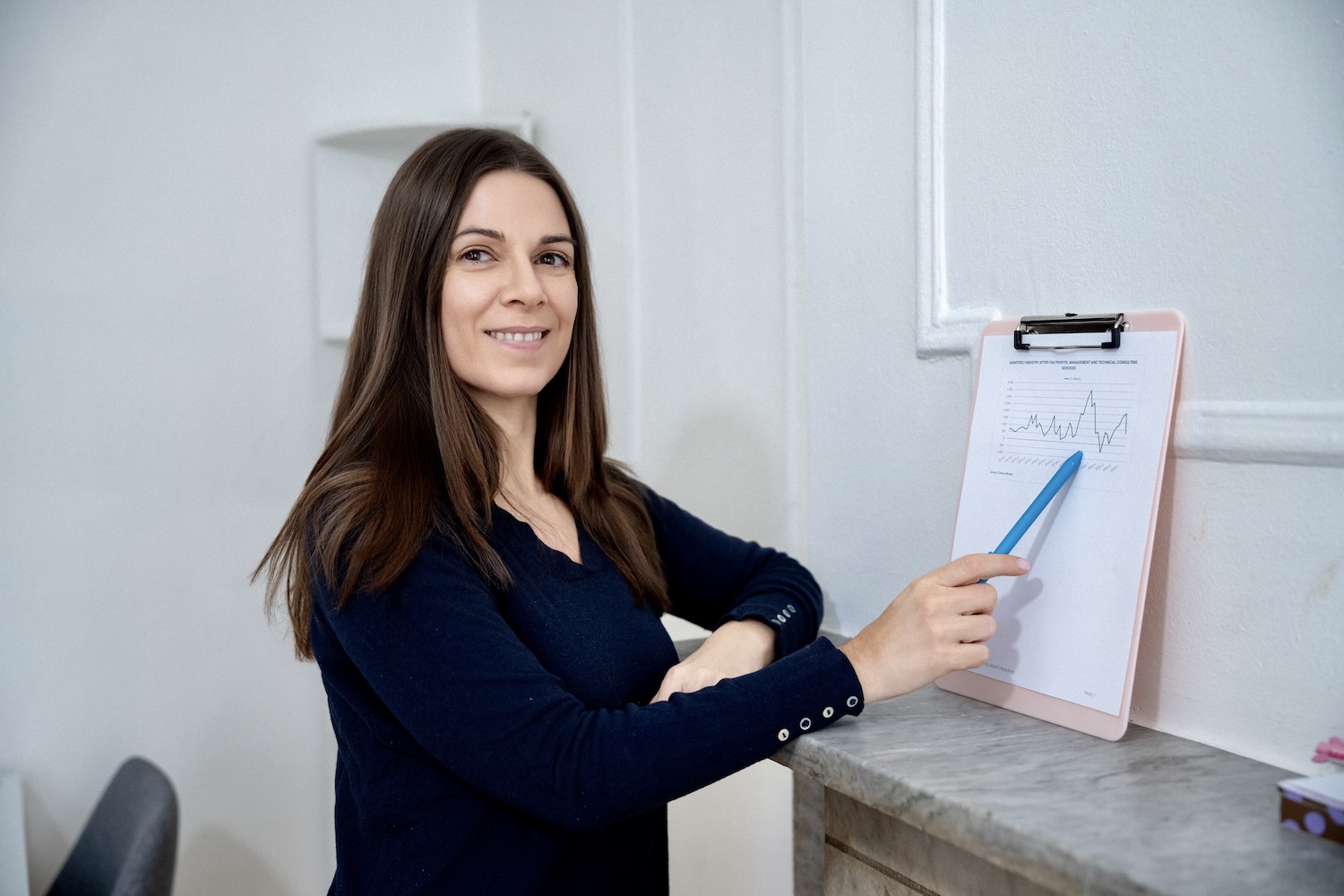 Woman in navy sweater holding clipboard with graph and pointing at it with a blue pen.
